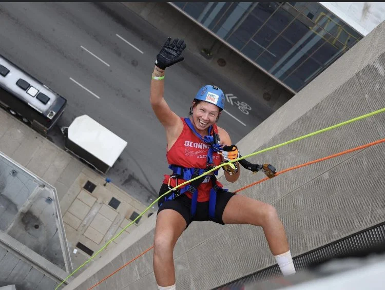 A woman rappelling down the side of a tall building, smiling and waving at the camera, wearing a helmet, gloves, and climbing gear.