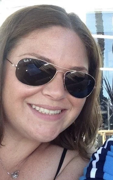 A smiling woman with brown hair wearing Ray-Ban aviator sunglasses and a black strap top, outdoors in a sunny setting.