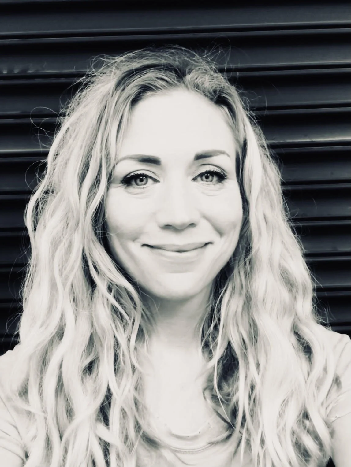 A woman with wavy blonde hair smiling at the camera, standing in front of dark horizontal blinds.