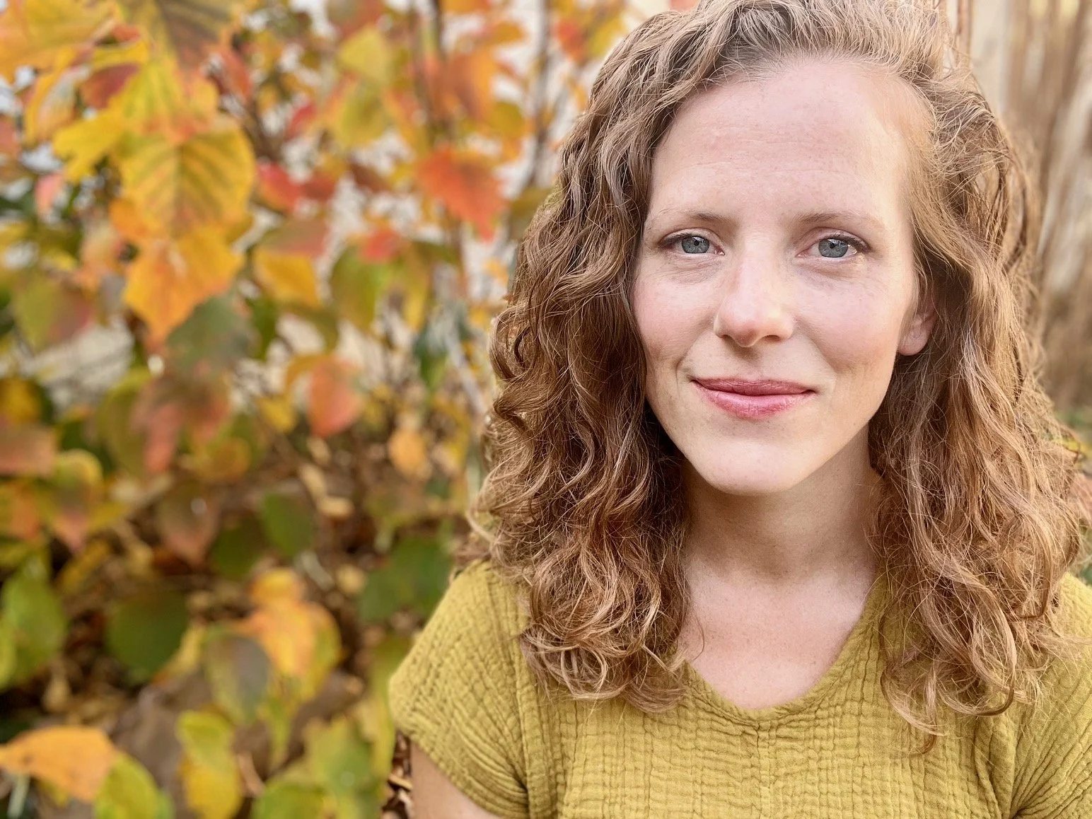 Close-up portrait of a young woman with curly red hair and blue eyes, smiling softly, outdoors with autumn-colored leaves in the background.