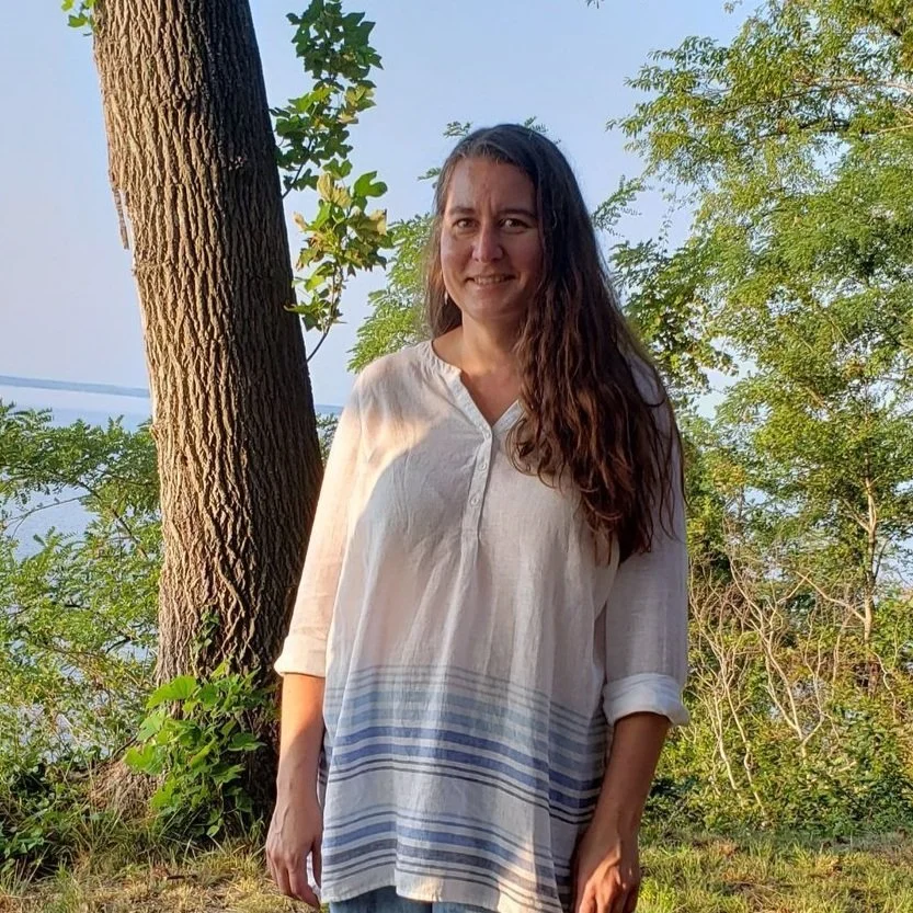 A woman with long, wavy brown hair wearing a white linen shirt with blue horizontal stripes at the hem, standing outdoors near a tree with green foliage in the background.