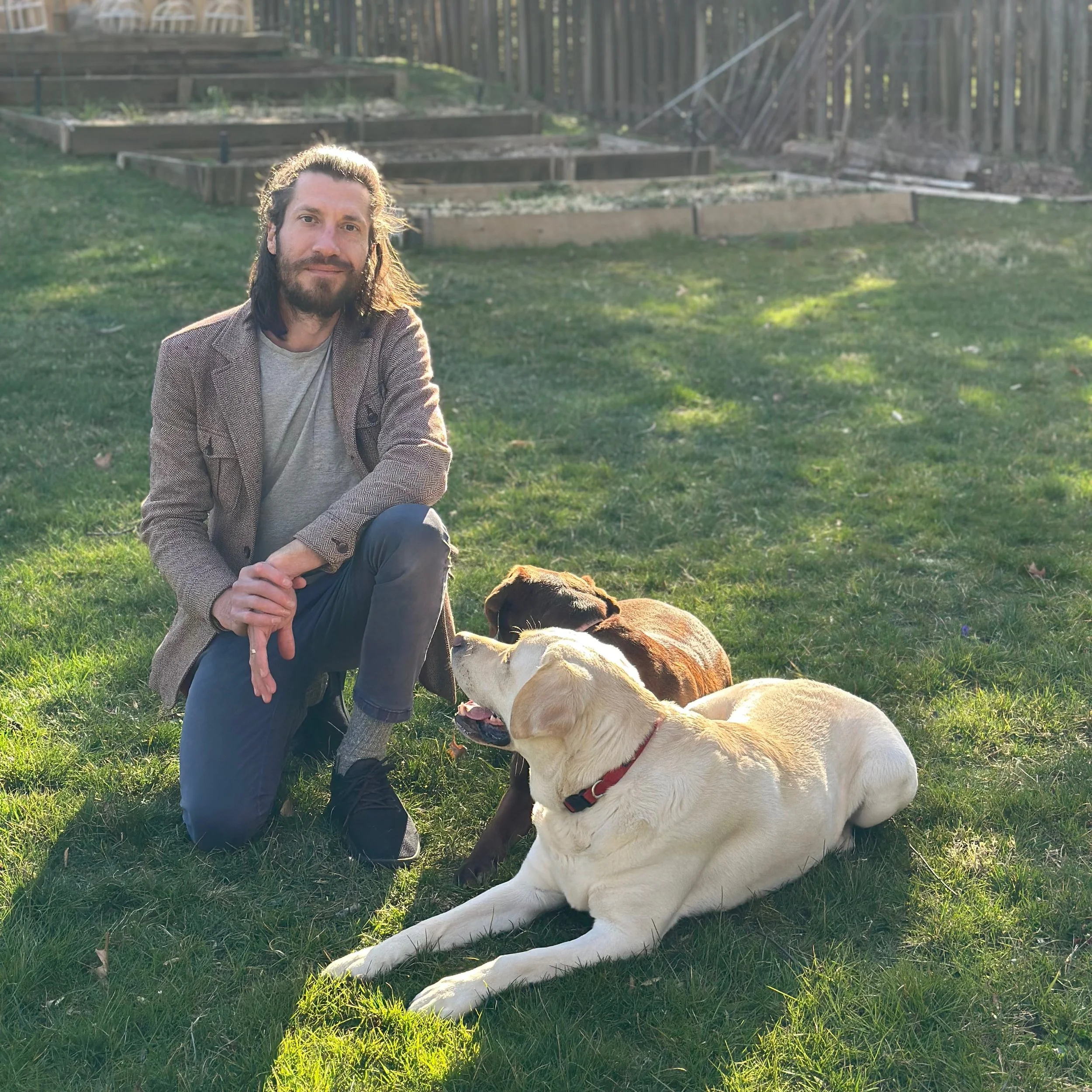 A man with long hair and a beard kneeling on grass with three dogs, two laying down and one sitting, in a backyard with wooden raised garden beds and a fence in the background during sunlight.
