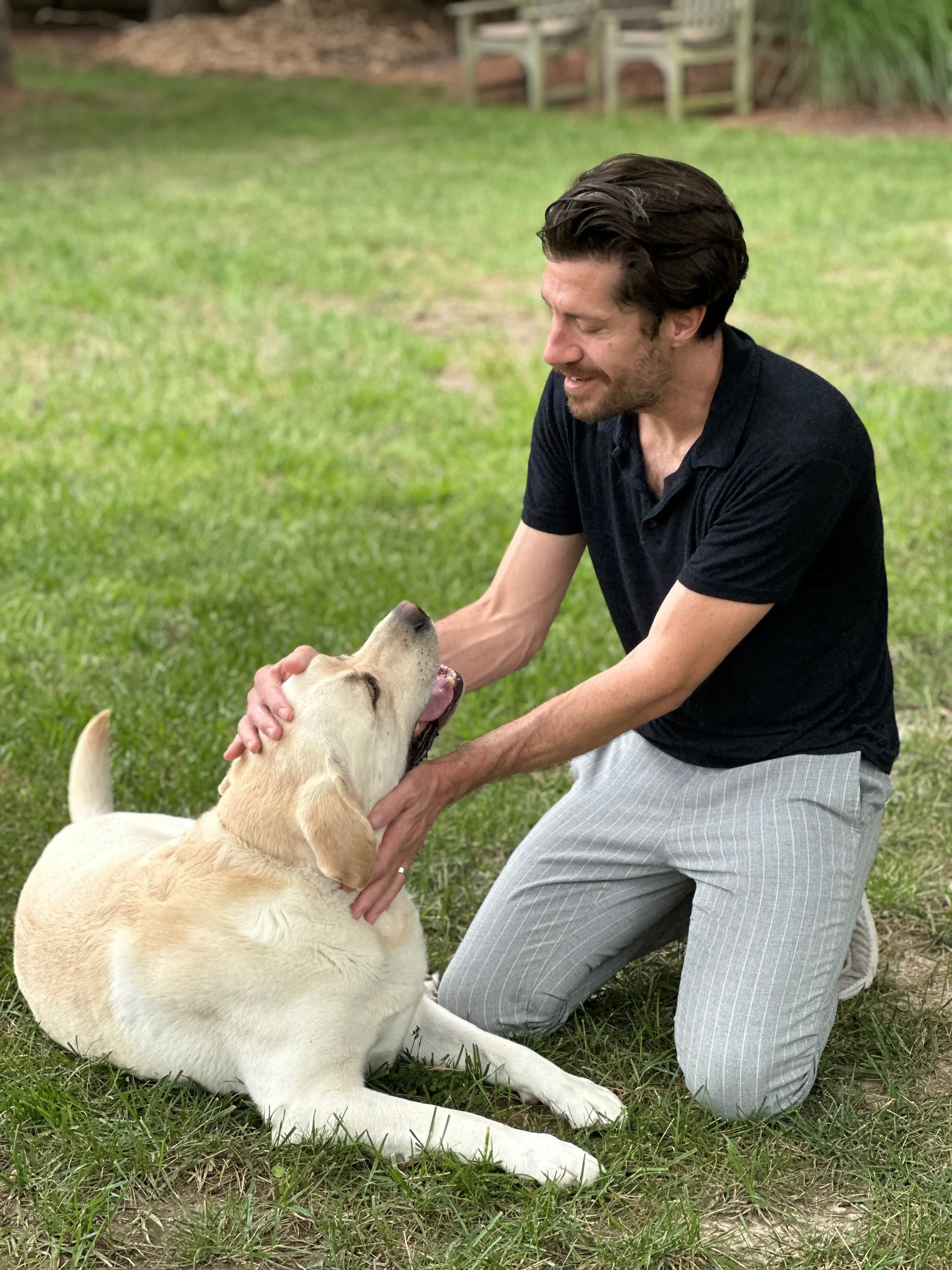 A man kneeling on the grass and petting a yellow Labrador retriever, both appearing happy and affectionate.