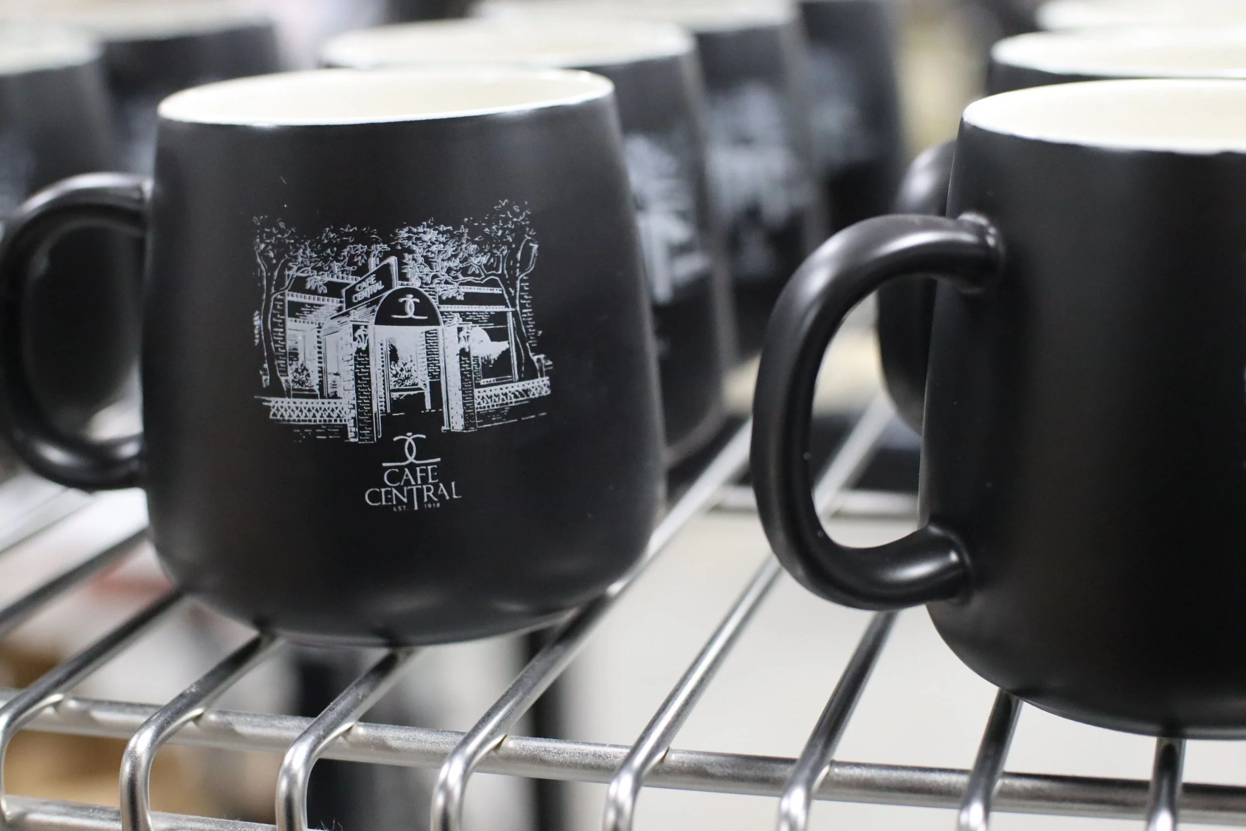 Black coffee mugs with a white logo reading "Cafe Central" on a metal wire shelf.