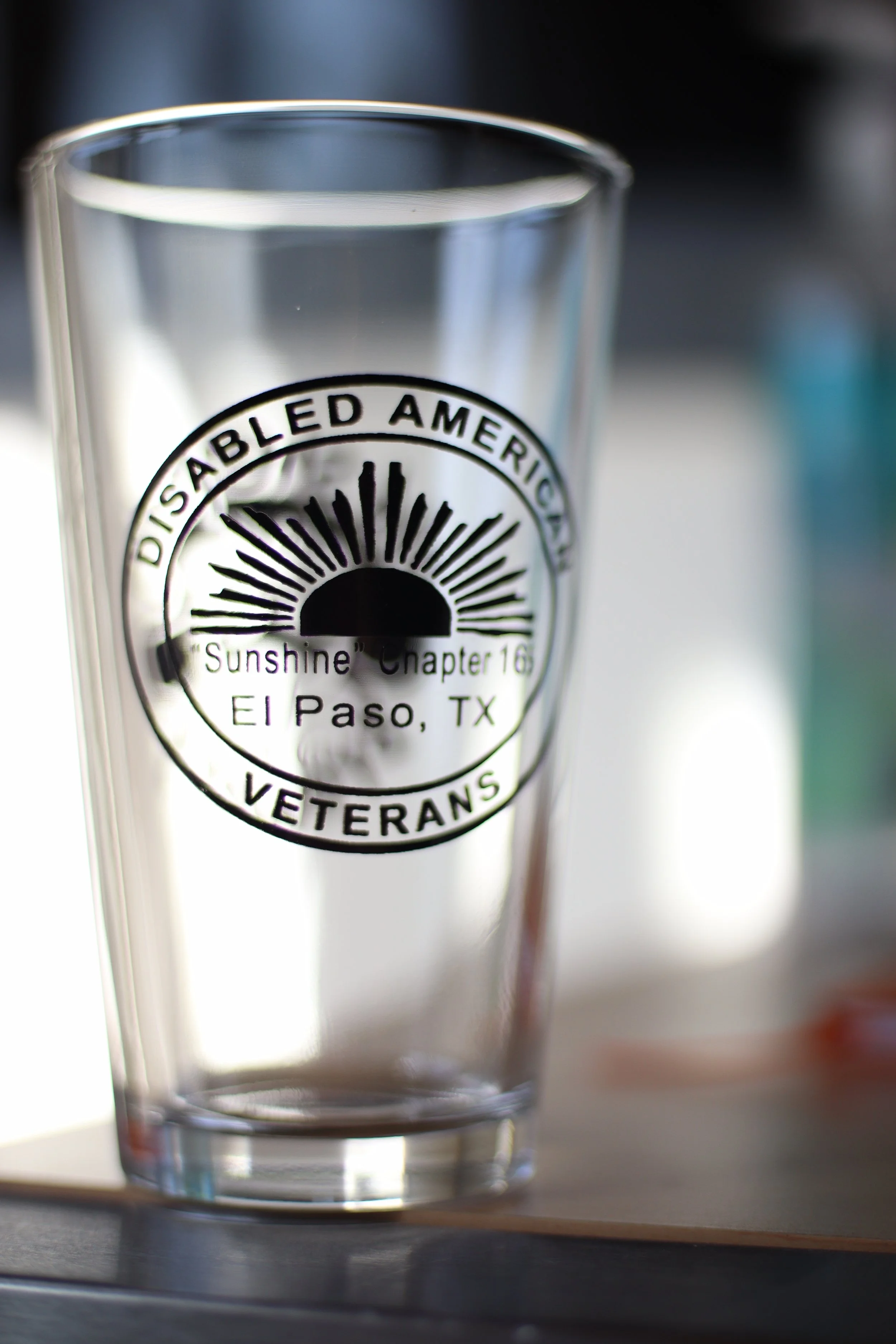 A clear Veterans Glass with a black logo that reads 'Disabled American Veterans, Sunshine Chapter 16, El Paso, TX.' The glass has a sunburst design in the logo. It is placed on a wooden surface with a blurred background.