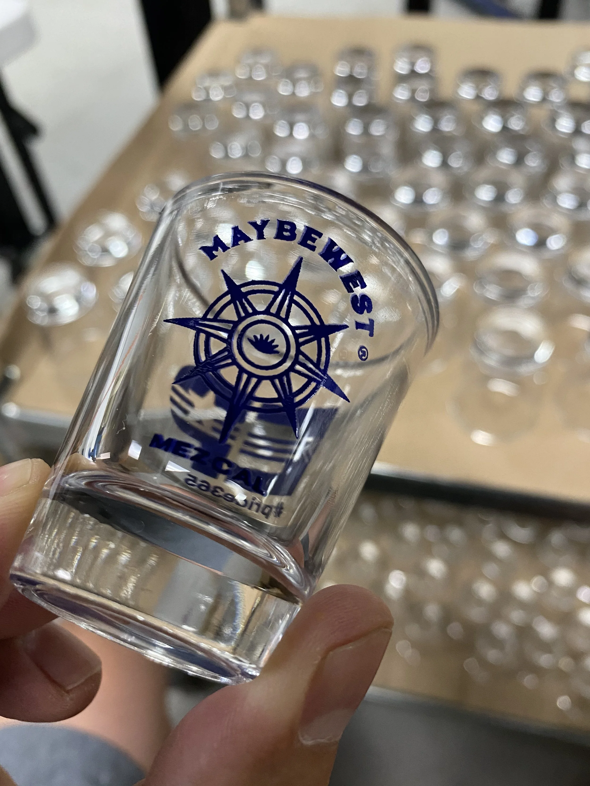 Close-up of a clear glass with blue text and a compass logo, held by a person's hand, with a background of multiple similar glasses and a wooden surface.