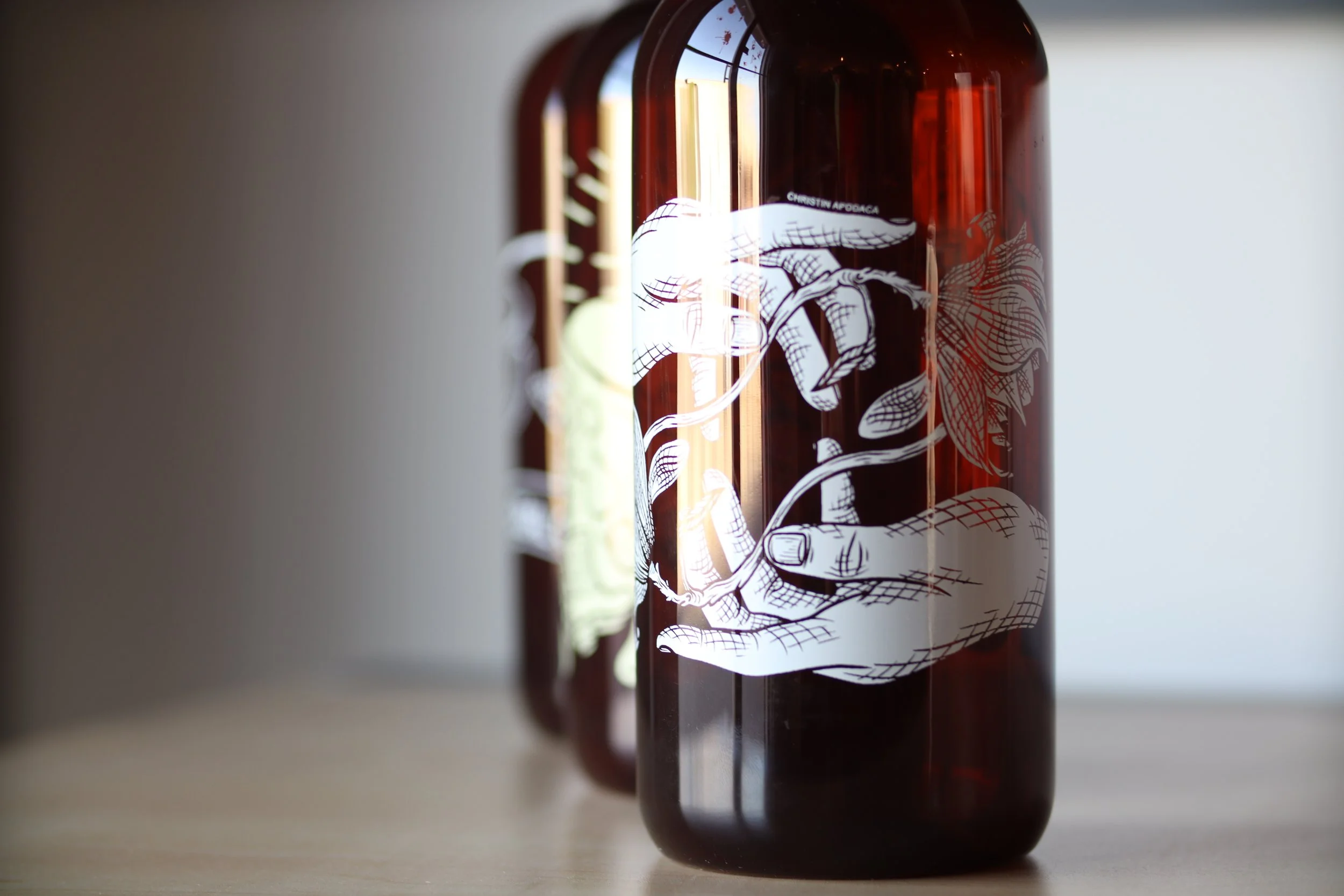 Custom-printed amber glass bottle featuring an intricate illustration of hands holding a plant, designed by MOAB for branded drinkware and breweries in El Paso, TX.