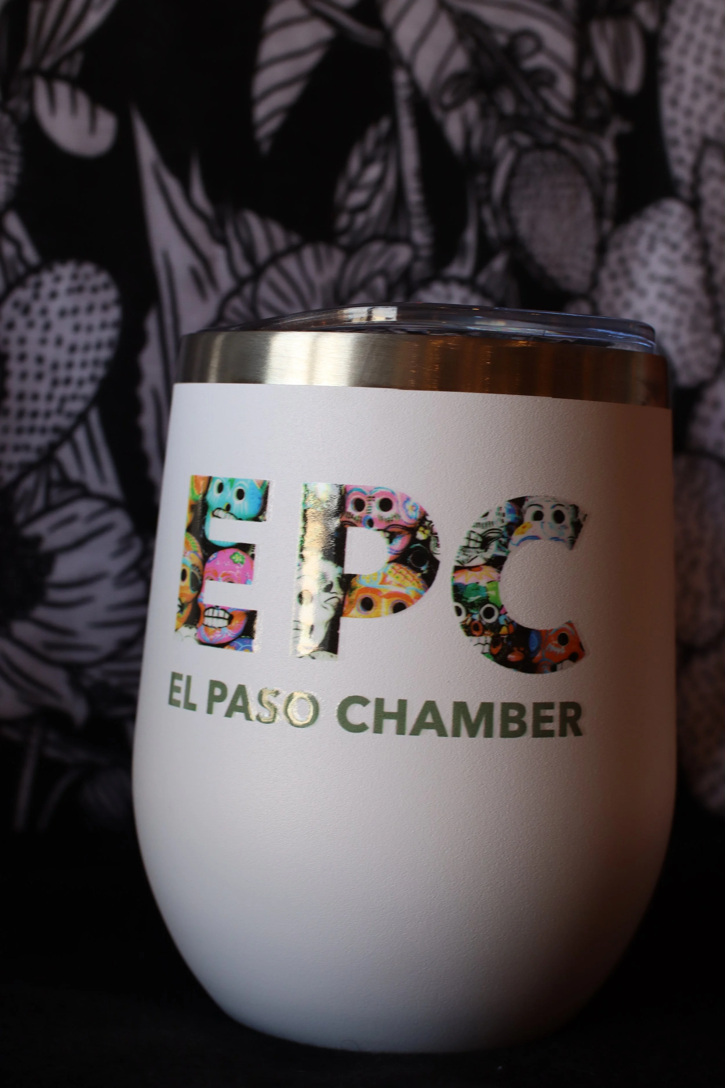 Close-up of a white beverage can with colorful, patterned lettering that reads "EPC" and additional text "El Paso Chamber," set against a dark, patterned fabric background.