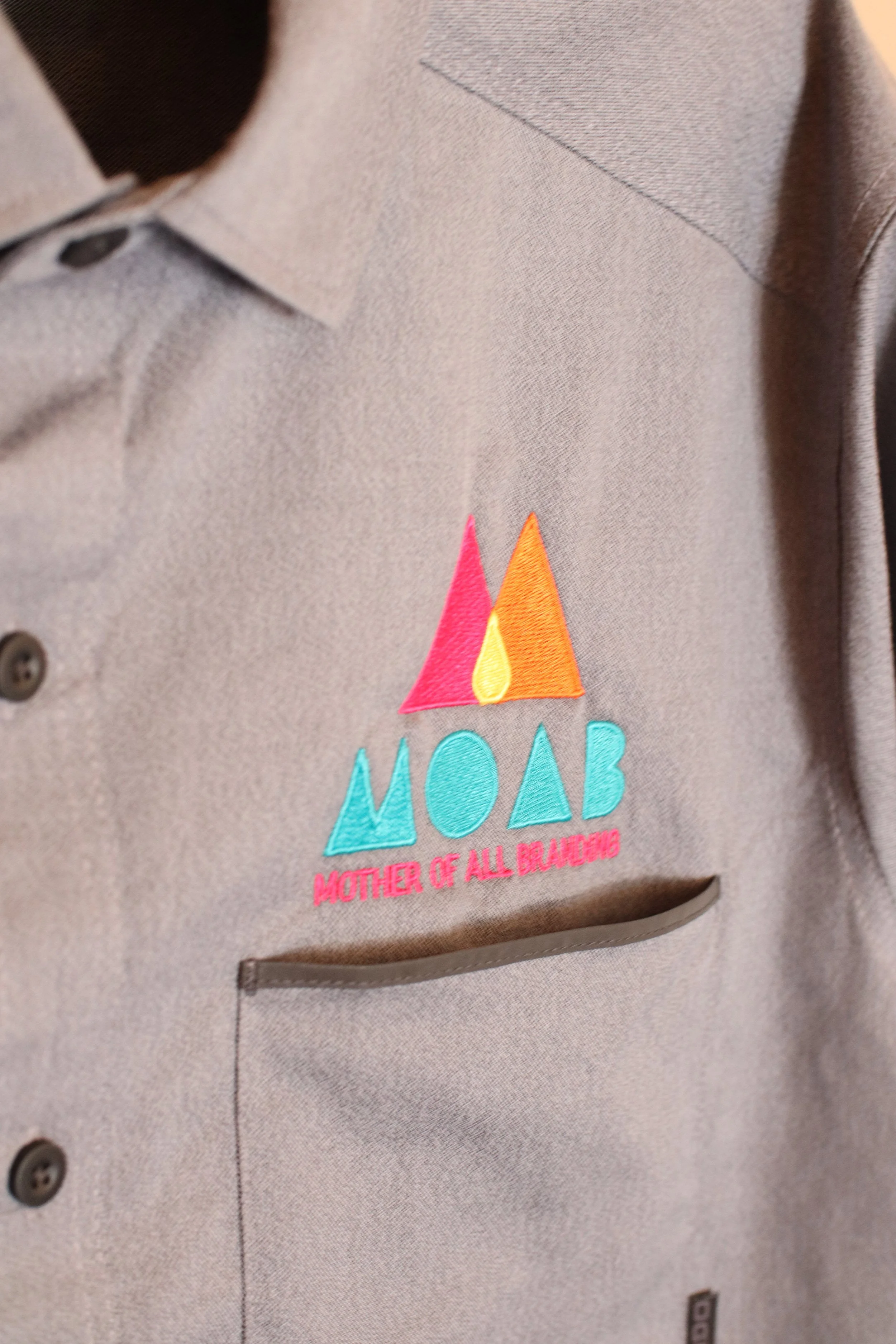 Custom-branded gray shirt with embroidered MOAB logo and "Mother of All Branding" text, designed for corporate apparel and work uniforms in Texas.