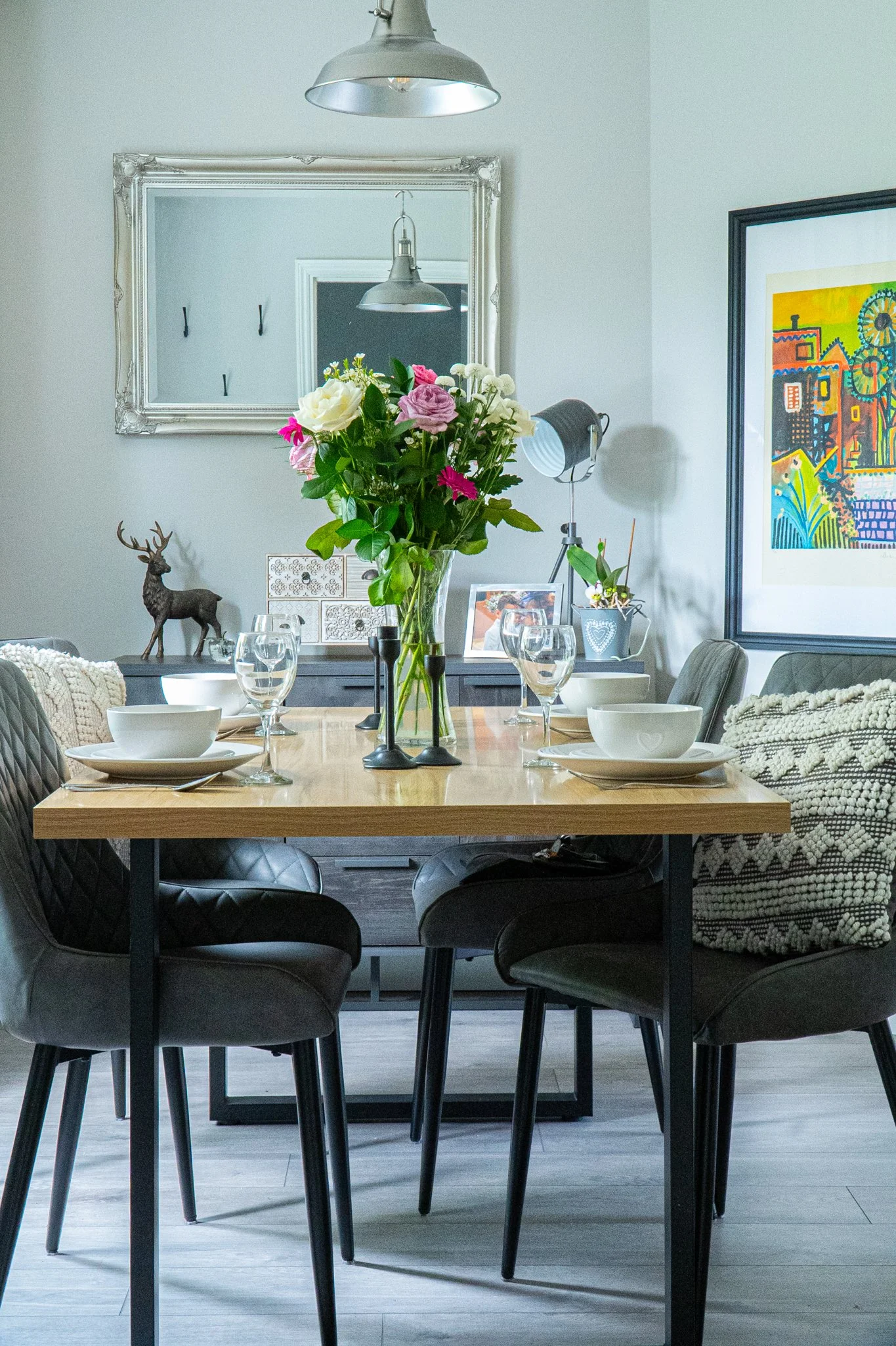 A dining room with a wooden table set with white bowls and glasses, surrounded by dark gray upholstered chairs, a large vase of mixed pink and white flowers, a mirror on the wall, and colorful artwork.