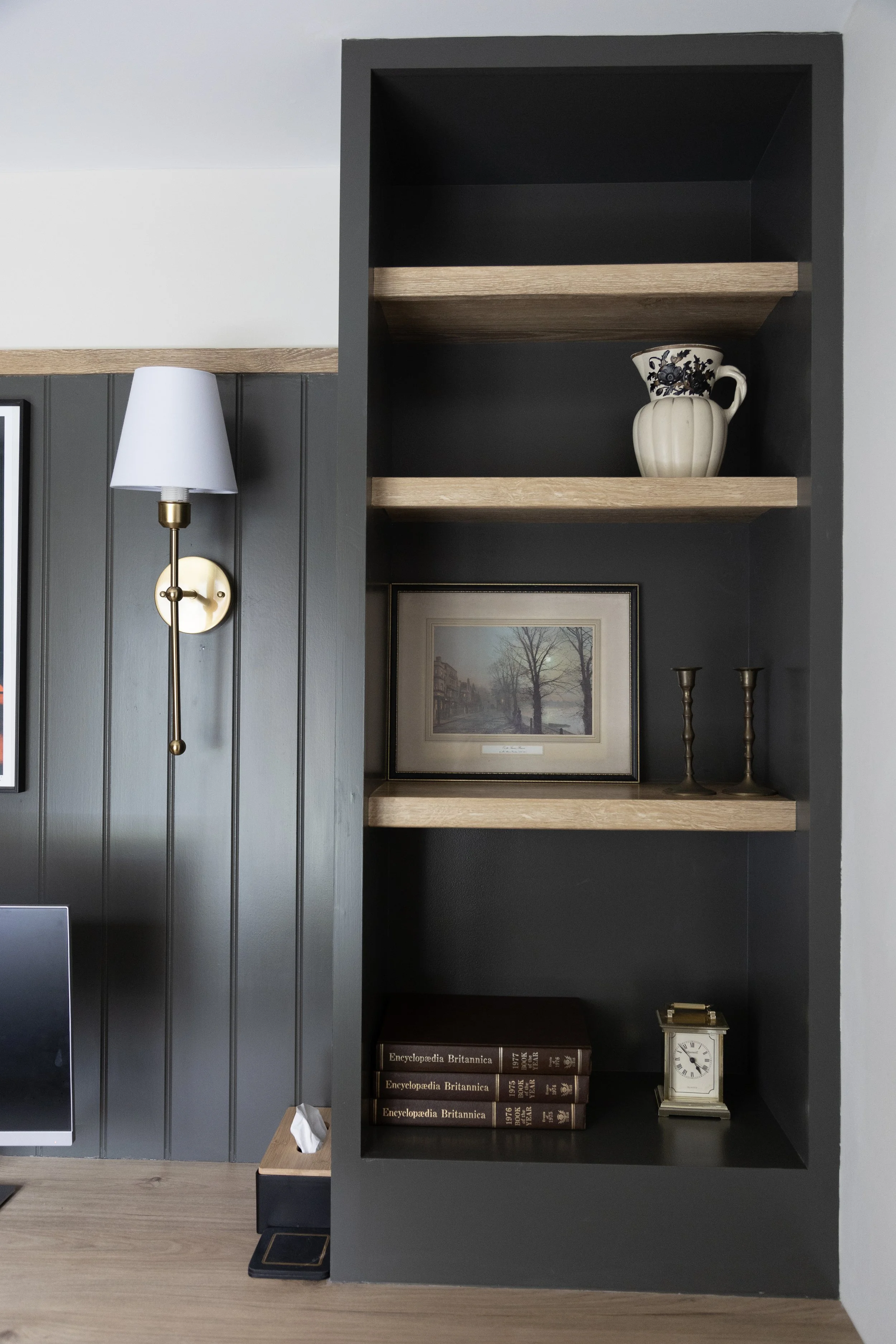 Interior bookshelf with a decorative pitcher on the middle shelf, framed artwork, and a stack of encyclopedias on the bottom shelf. To the left, a wall-mounted lamp and a tissue box are visible.