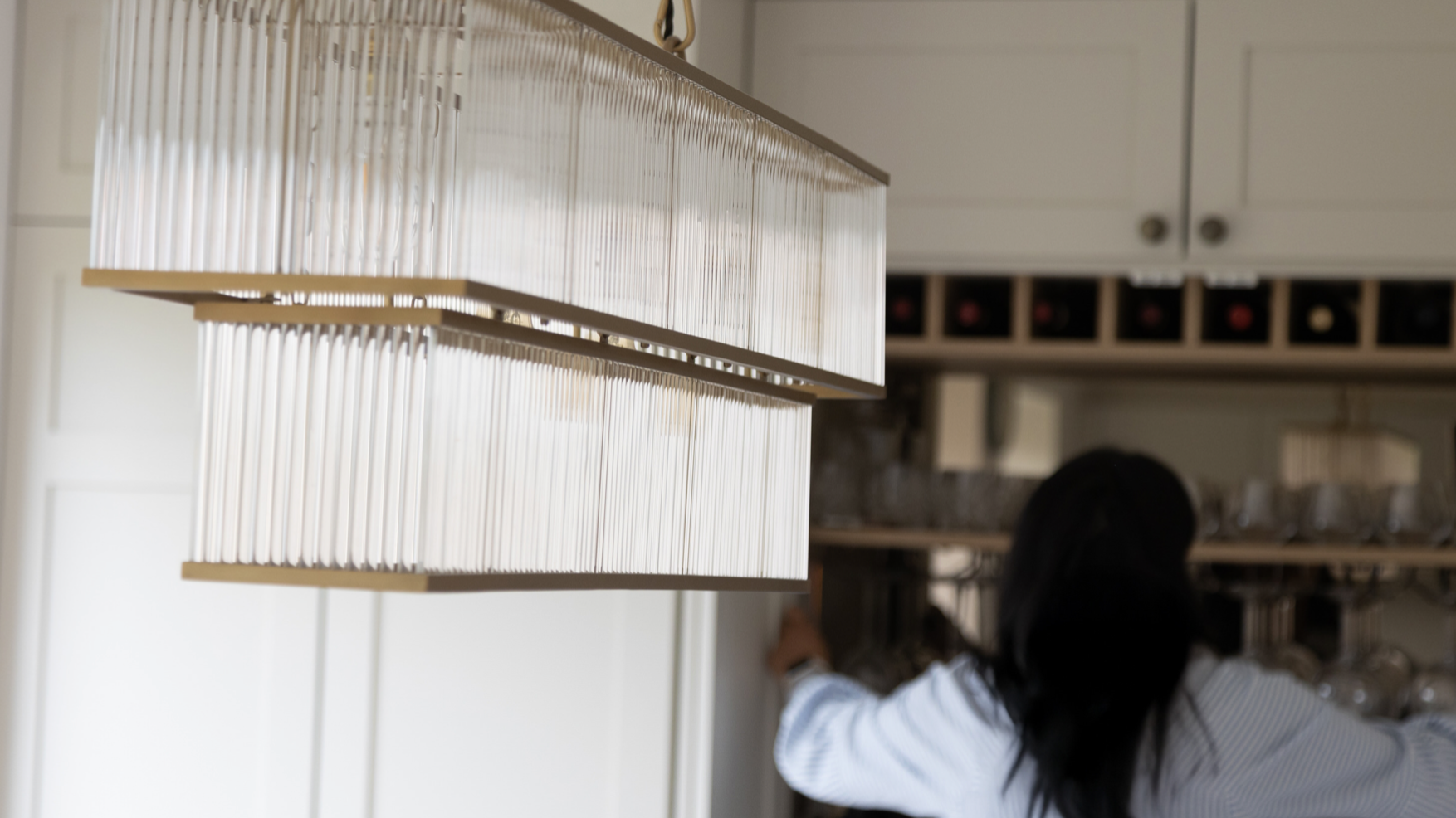 Close-up of a hanging transparent light fixture in a kitchen, with a woman reaching for items on a shelf in the background.