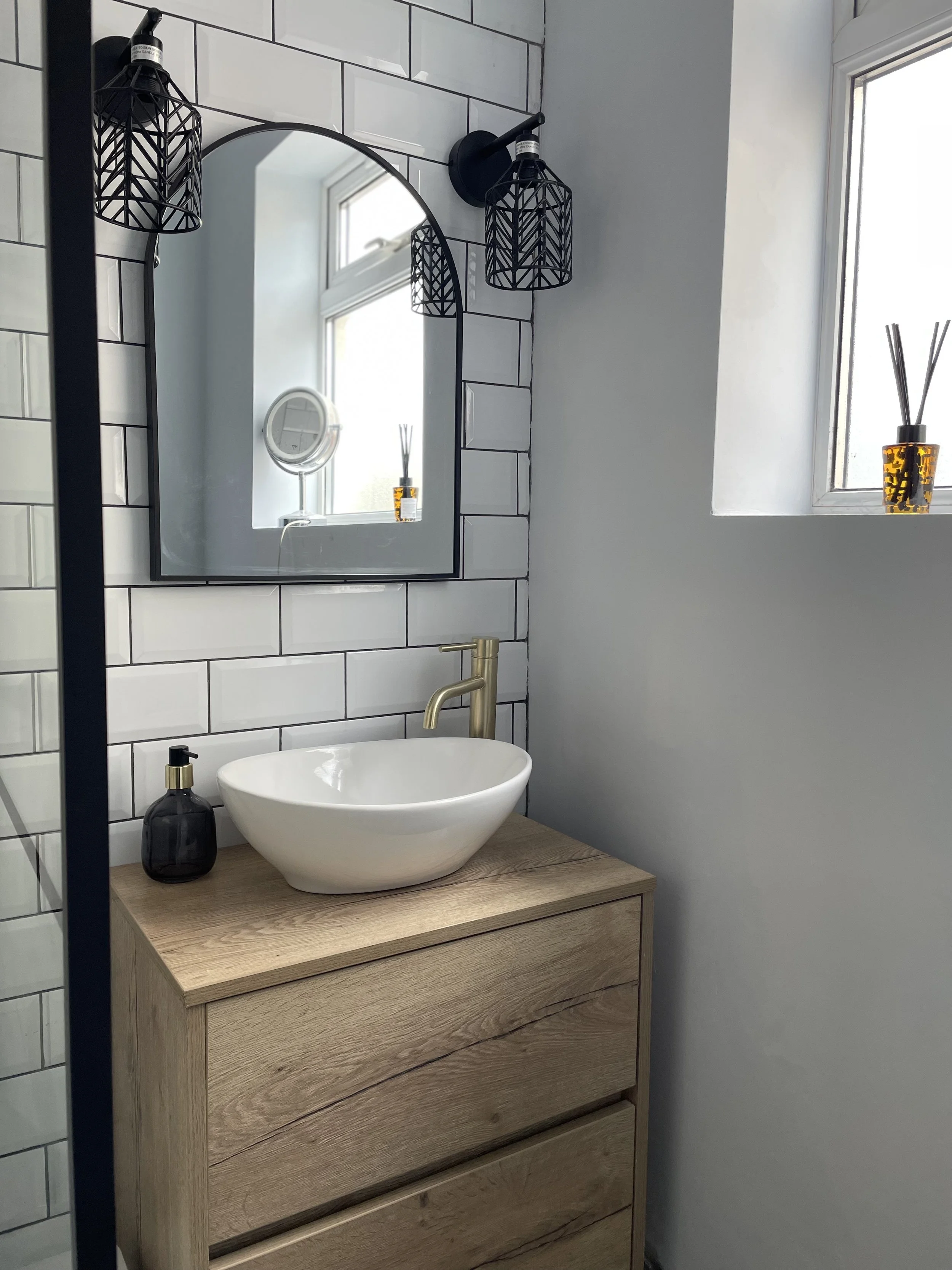 Modern bathroom with a wooden vanity, white vessel sink, gold faucet, large wall mirror, black soap dispenser, black wall sconces, and a window with a small decorative plant in a yellow and black pot.