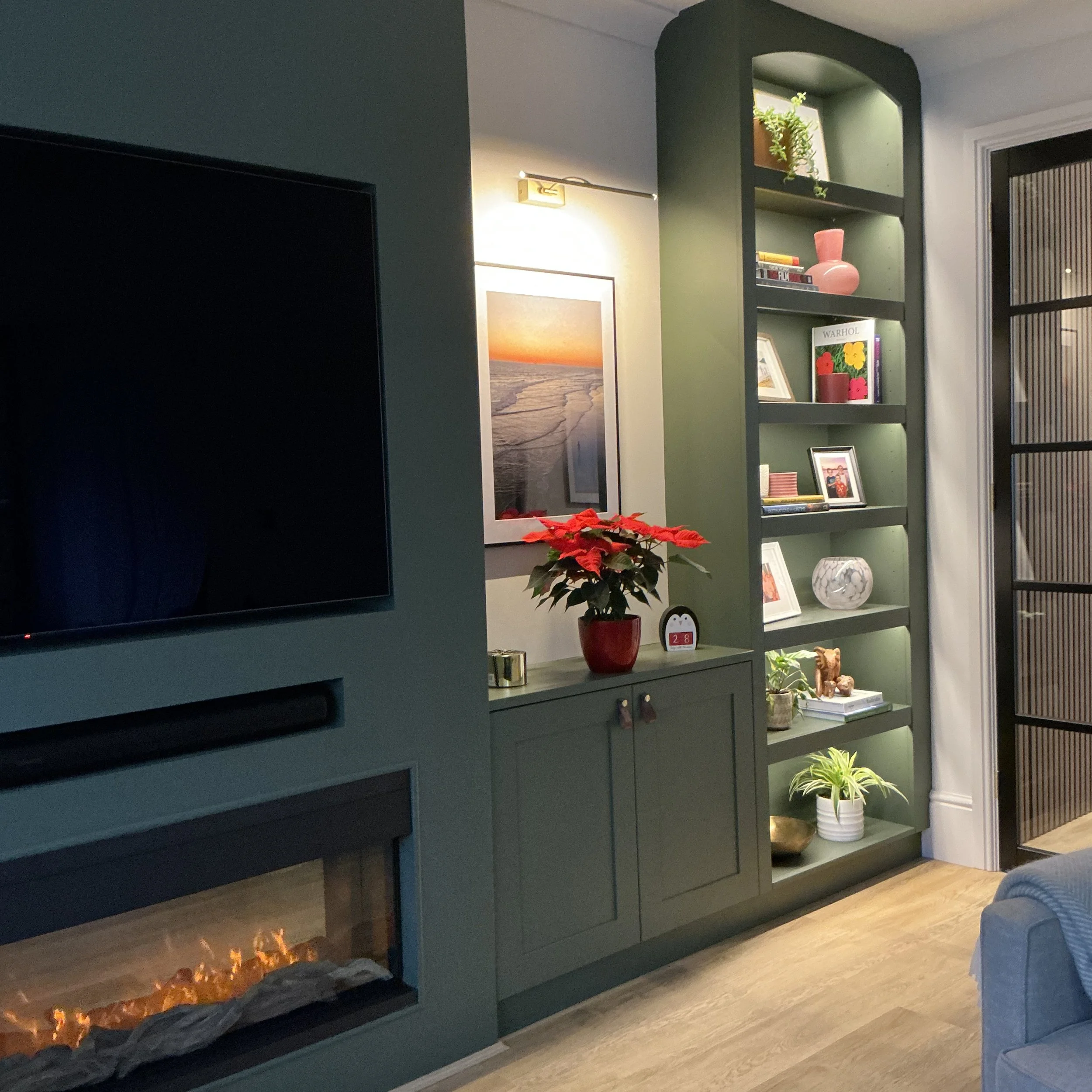 Living room with a dark blue fireplace and a teal bookcase filled with books, decorative items, and framed photos. A plant in a red pot sits on the TV stand, and a poinsettia plant is nearby. The room has a wooden floor, and a window with curtains is