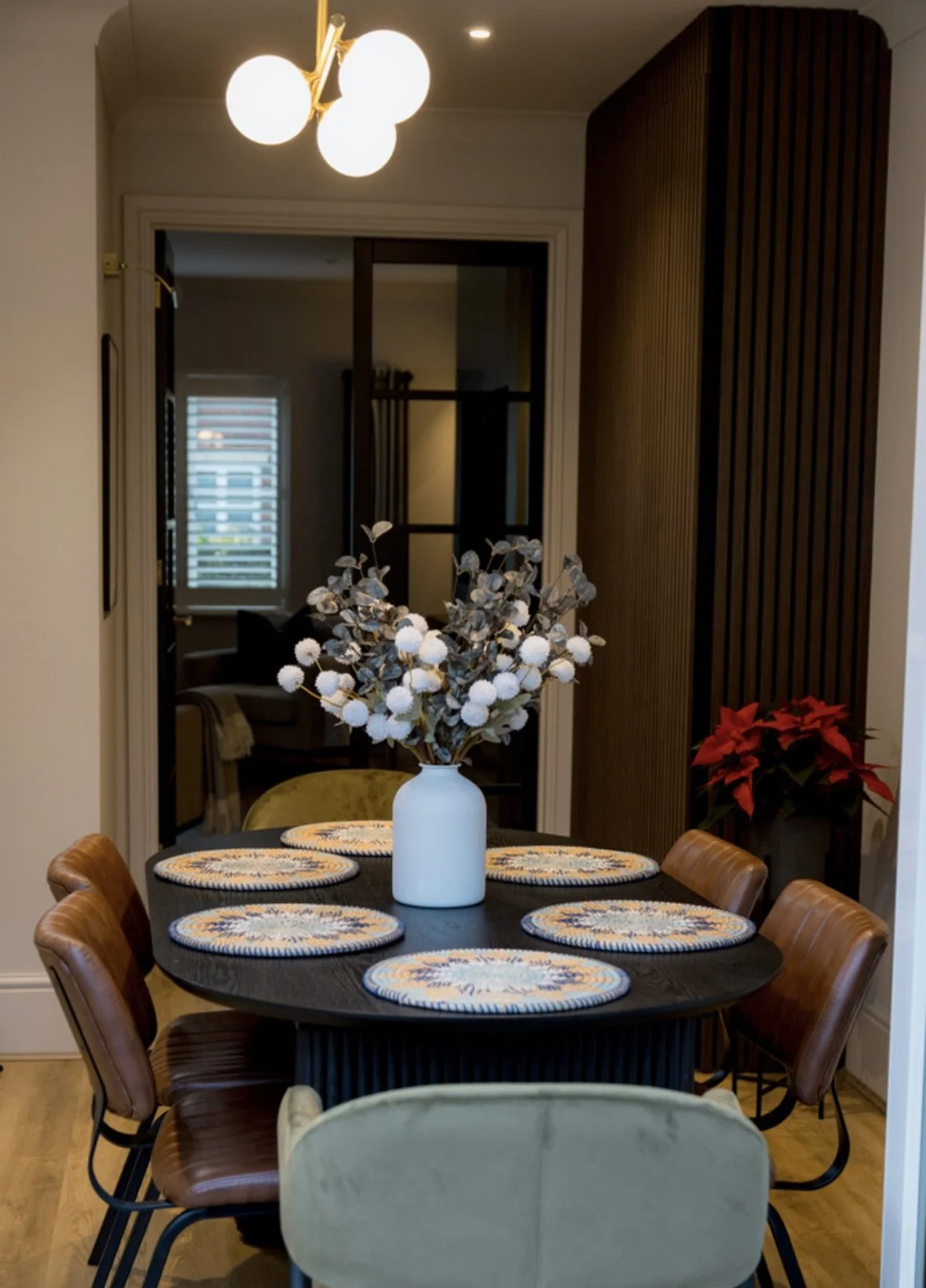 A dining room with a black round table decorated with six woven placemats and a white vase with white floral arrangements. Six brown chairs are around the table, and a beige chair is in the foreground. There is a window with shutters, and a red poins