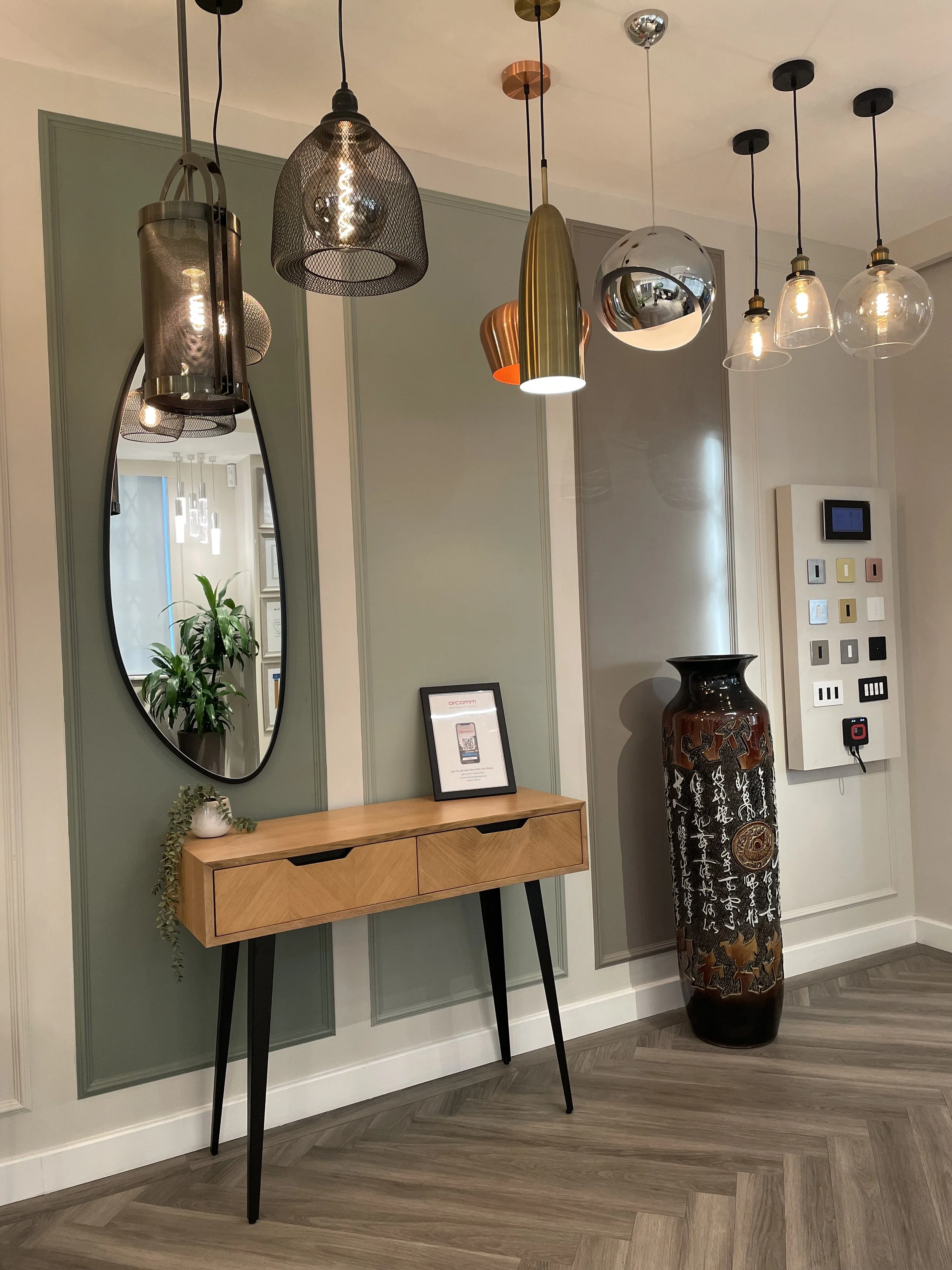 A modern interior with a mirror, hanging pendant lights, a small wooden console table with black legs, a framed sign, a decorative plant, and a tall vase with Asian script and animal motifs, against a wall with green and gray paneling.