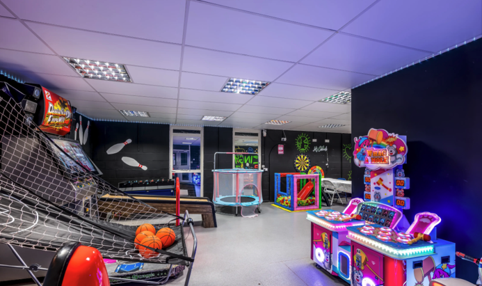 Indoor arcade with various gaming machines and entertainment options, including claw machines, basketball hoops, and digital games, on dark walls with ceiling lights.