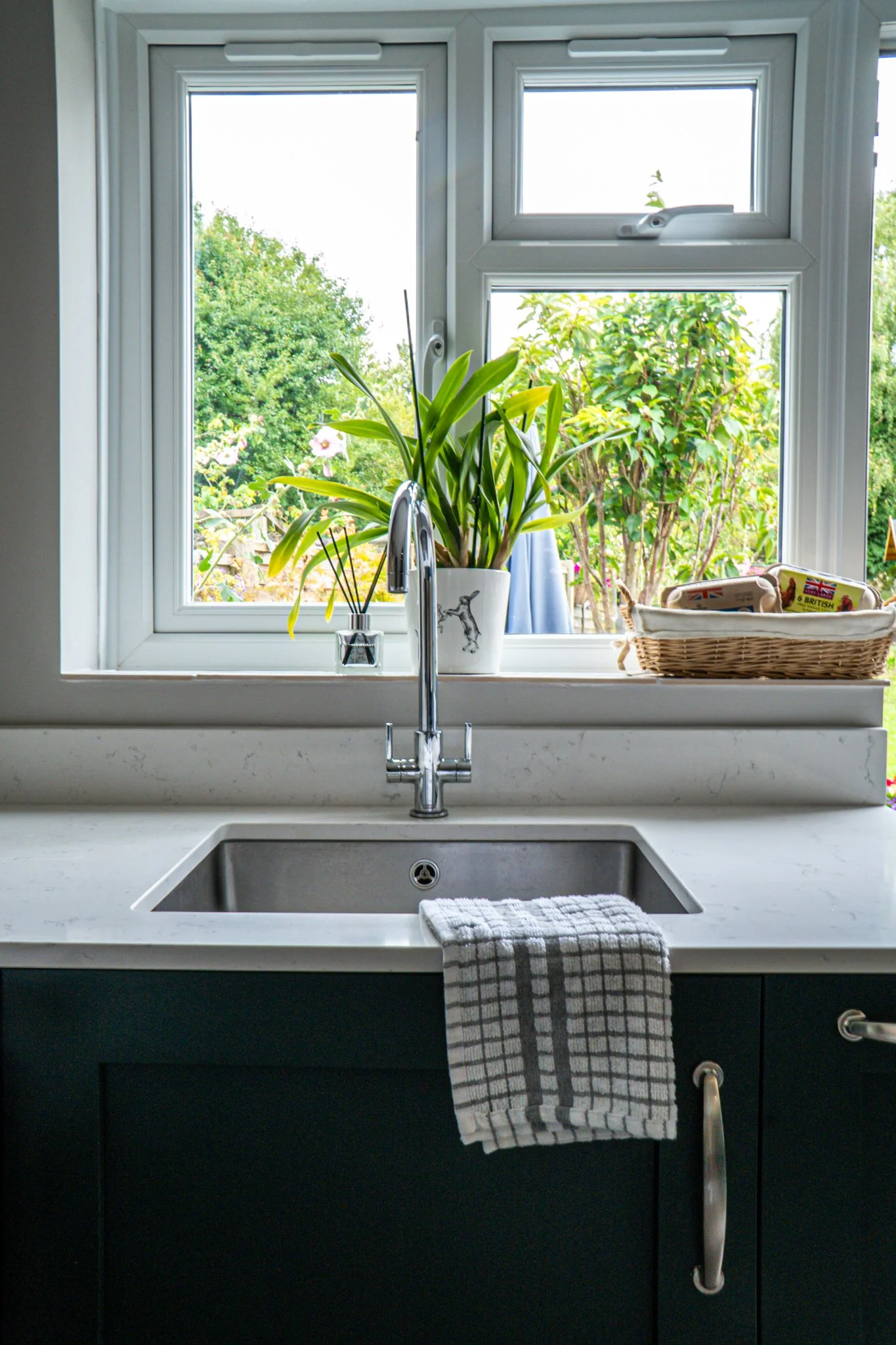 Kitchen sink with a striped dish towel, a potted plant, a basket, and a window showing greenery outside.