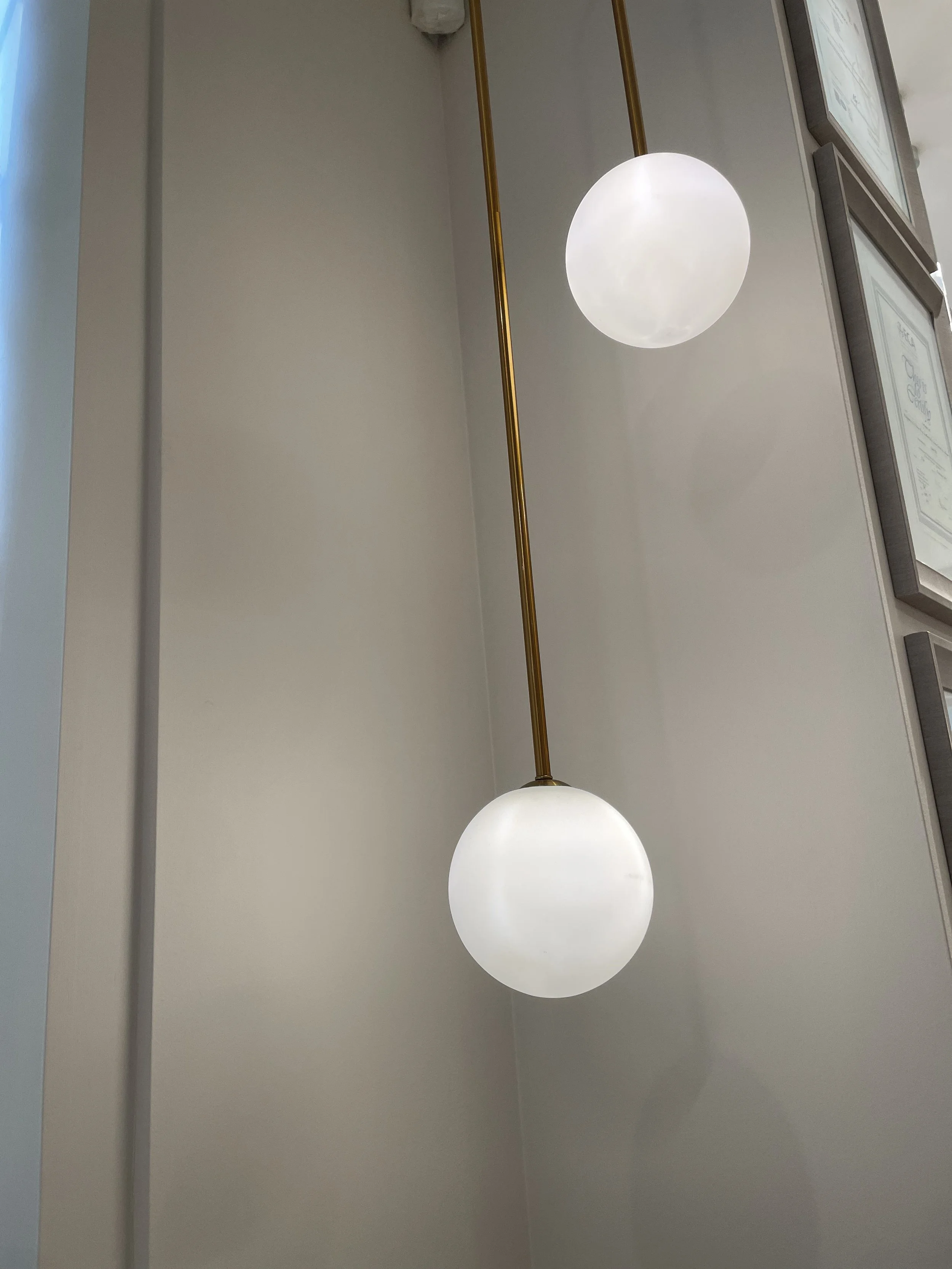A modern hanging spherical light fixture with a frosted glass globe, suspended from a brass rod attached to the ceiling, seen against a plain white wall in an indoor space.