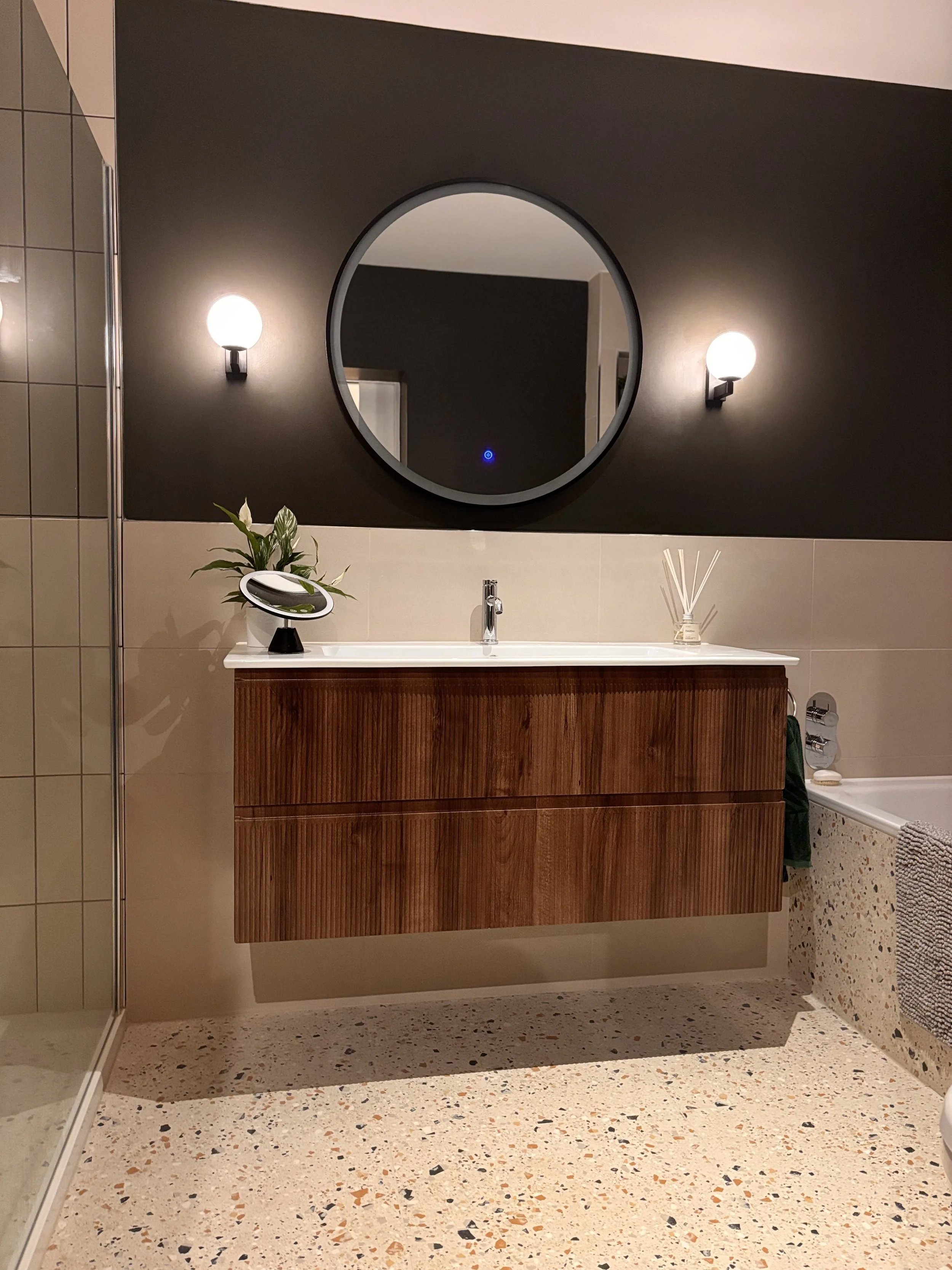 Bathroom vanity with a wooden cabinet, a white sink, a round mirror, and two wall sconces on a dark wall. A plant, a mirror, and reed diffusers are on the countertop. The bathroom has terrazzo flooring.