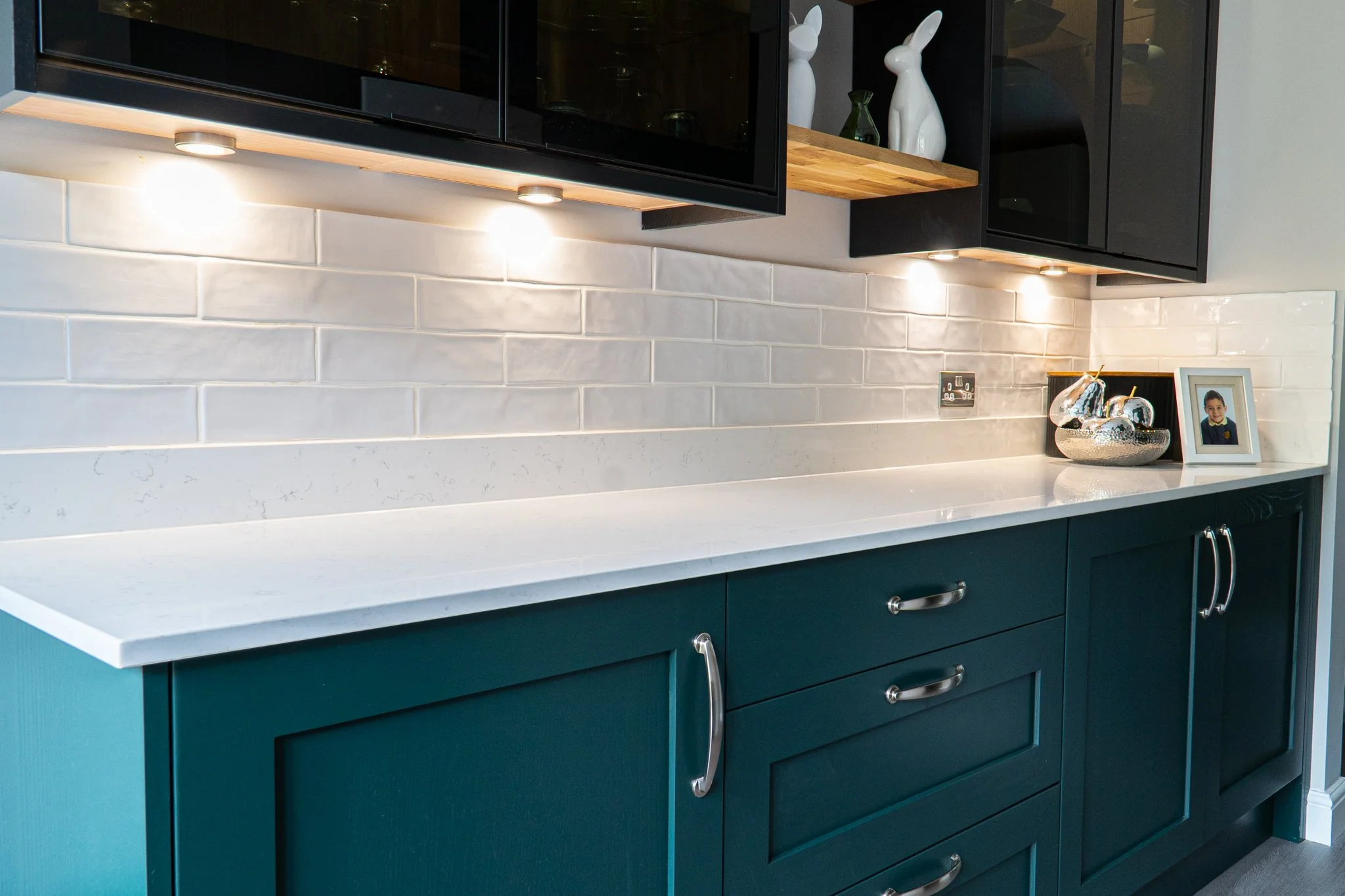 Close-up of a modern kitchen countertop with teal cabinets, silver handles, a white countertop, white subway tile backsplash, decorative rabbit-shaped vases, a framed photo of a boy, and a silver bowl with decorative ball ornaments.