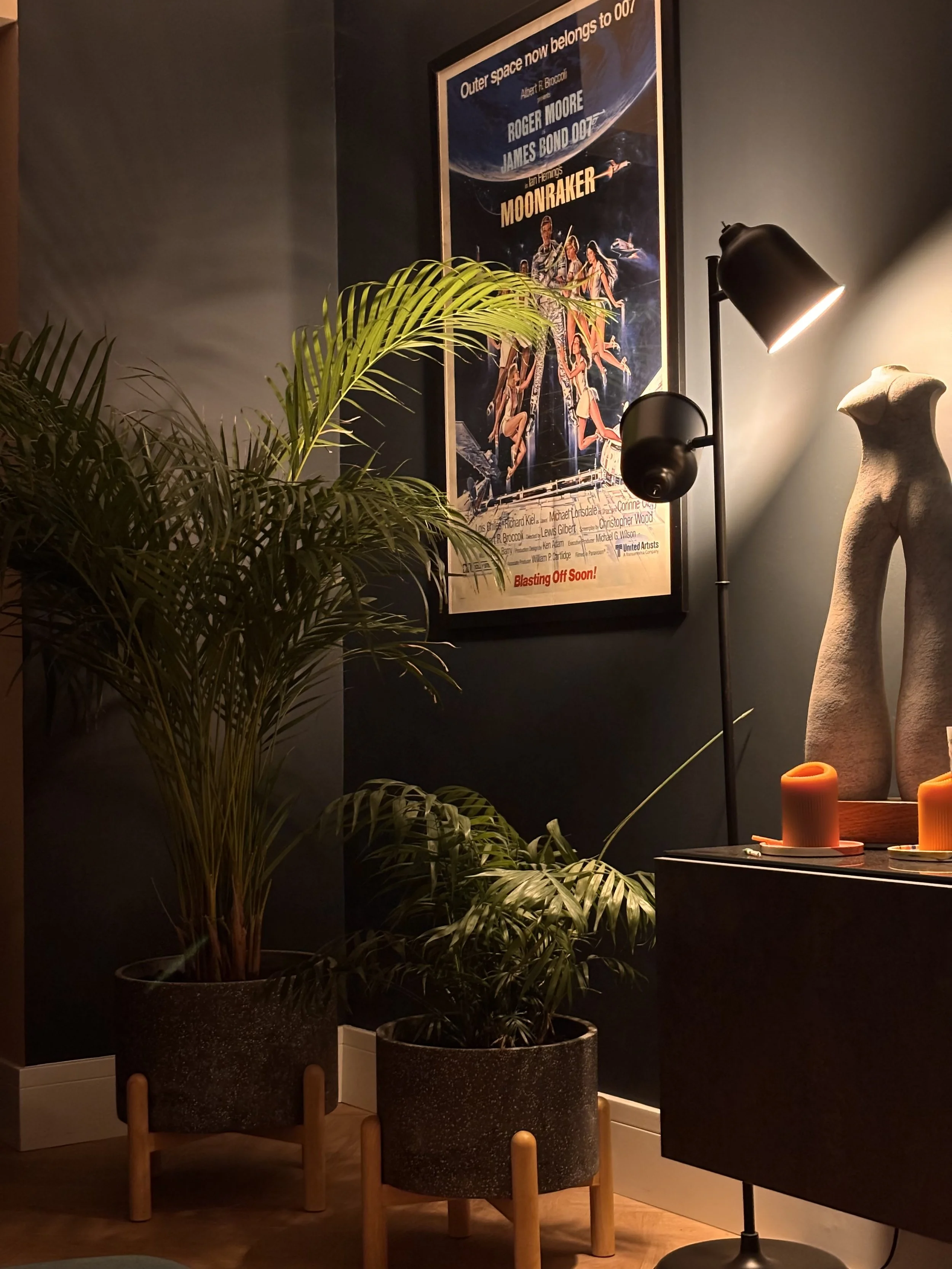A dimly lit interior space with a framed vintage James Bond movie poster on the dark wall, a tall green potted plant, a black floor lamp with a bright light, a sandstone statue of a torso, and orange candles on a side table.