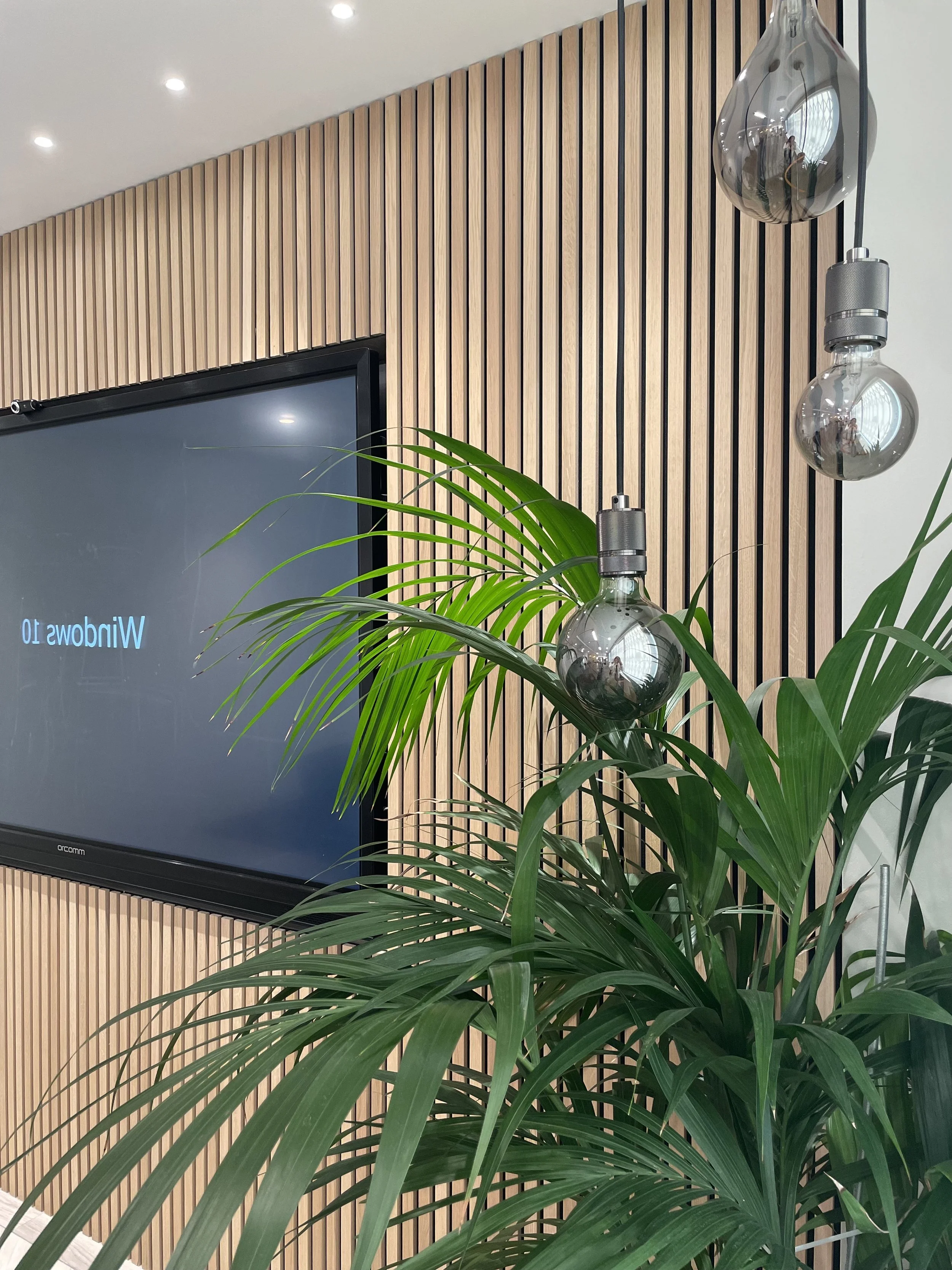 Interior space with a wooden slatted wall, a large flat screen TV, green potted plant, and hanging glass light fixtures.