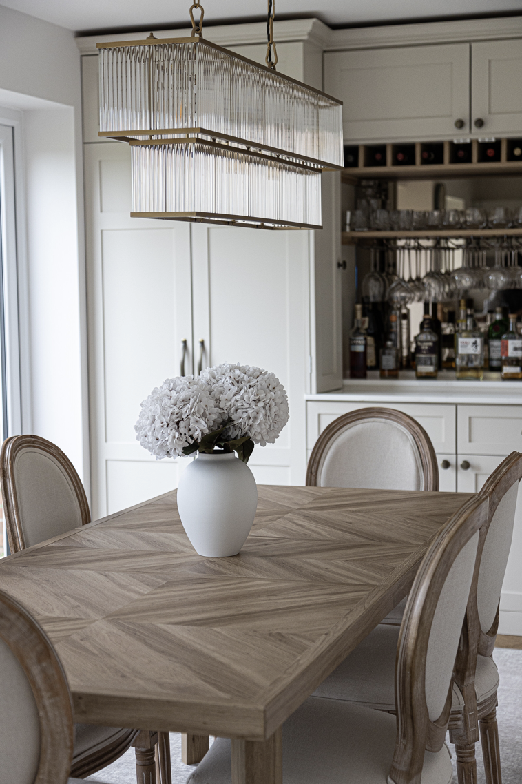 A dining room with a wooden table, beige upholstered chairs, a white vase with white flowers on the table, a modern rectangular chandelier above, and a wine and liquor cabinet in the background.