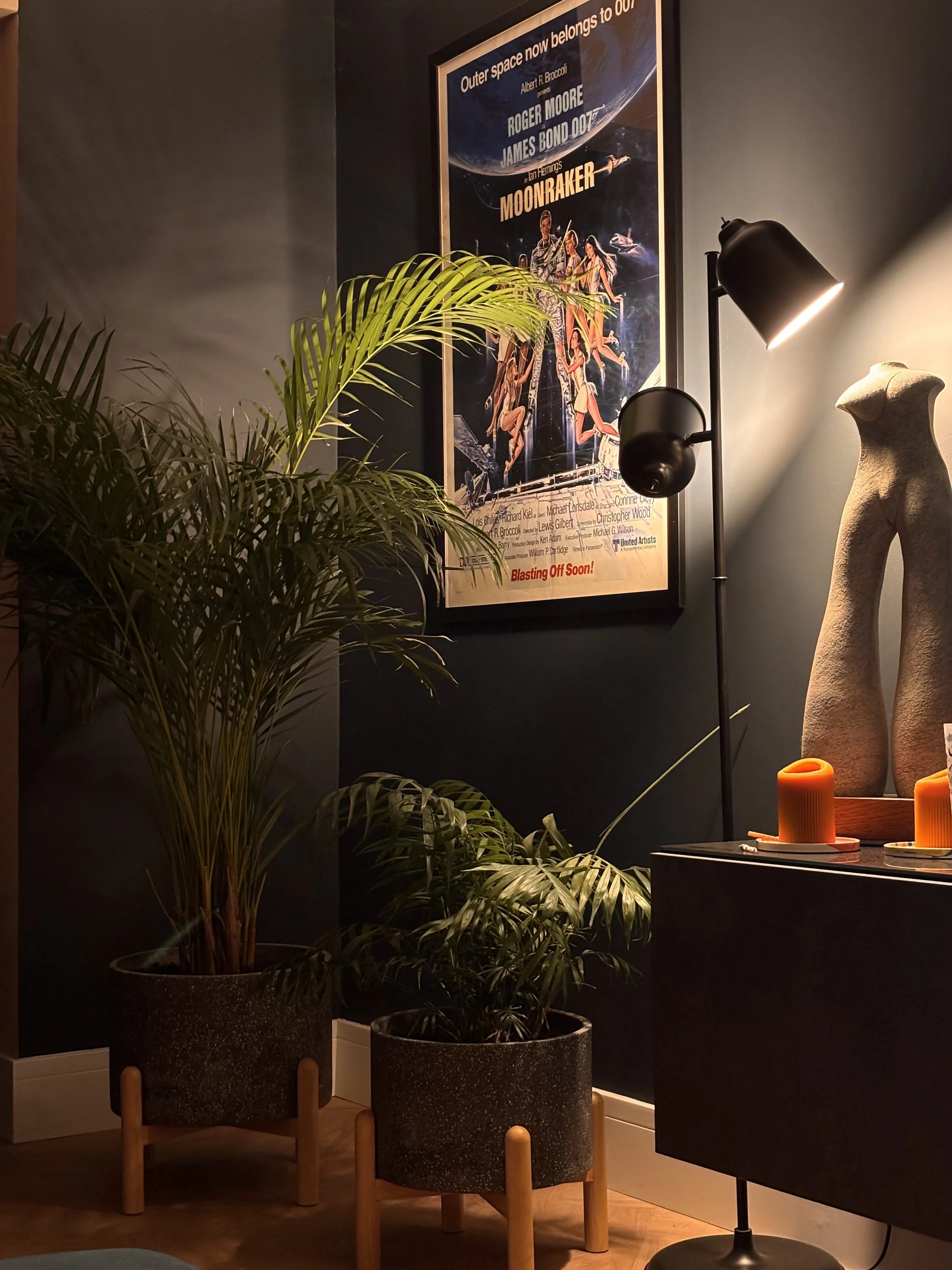 Indoor scene with two large potted plants, a modern black floor lamp, a presentation poster related to space, and a white mannequin torso on a black table with orange candles.