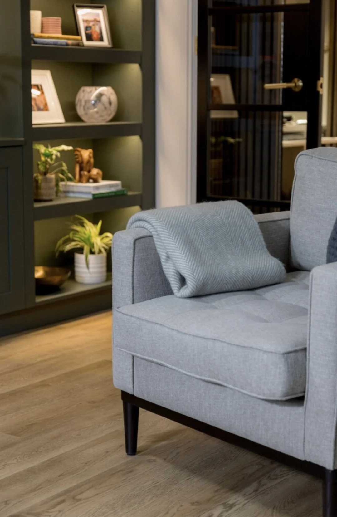 A light gray upholstered armchair with black wooden legs, a gray throw blanket draped over the armrest, and a plaid pillow on the seat. Behind the chair is a dark green bookshelf with decorative items, books, framed photos, a potted plant, and a marb