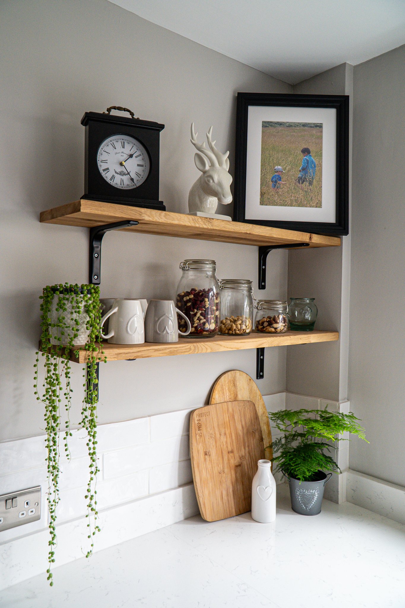 A kitchen with two wooden shelves on a light gray wall. The top shelf has a black clock, a white deer head sculpture, and a framed photo of two children in a field. The lower shelf holds glass jars with snacks, a potted trailing plant, and white mugs. A wooden cutting board leans against the wall, and a small white vase and a gray pot with a fern are on the white countertop.