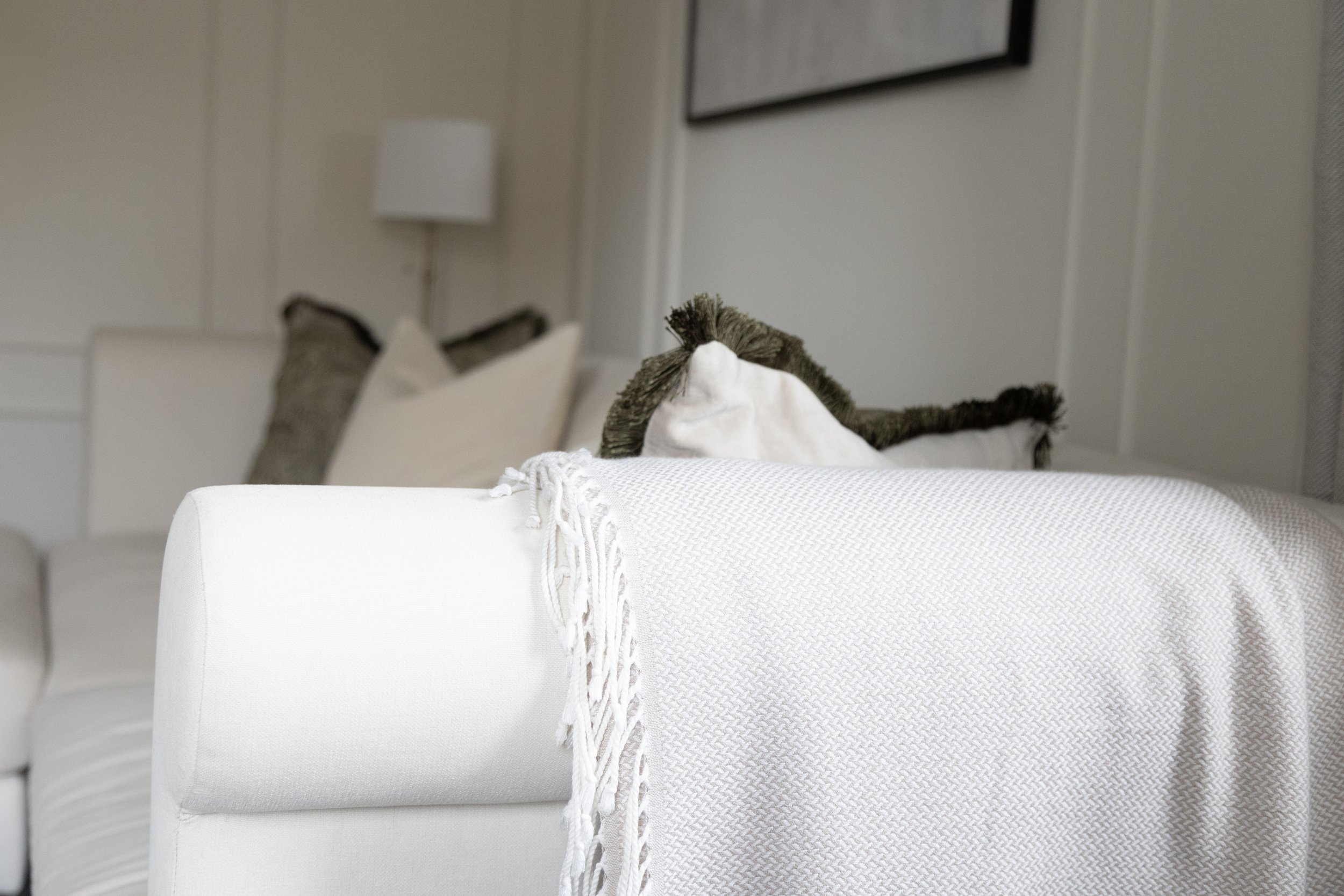 A close-up photo of a white textured blanket with tasseled edges draped over a piece of furniture in a living room. In the background, there are pillows on a sofa and picture frames on the cream-colored wall.