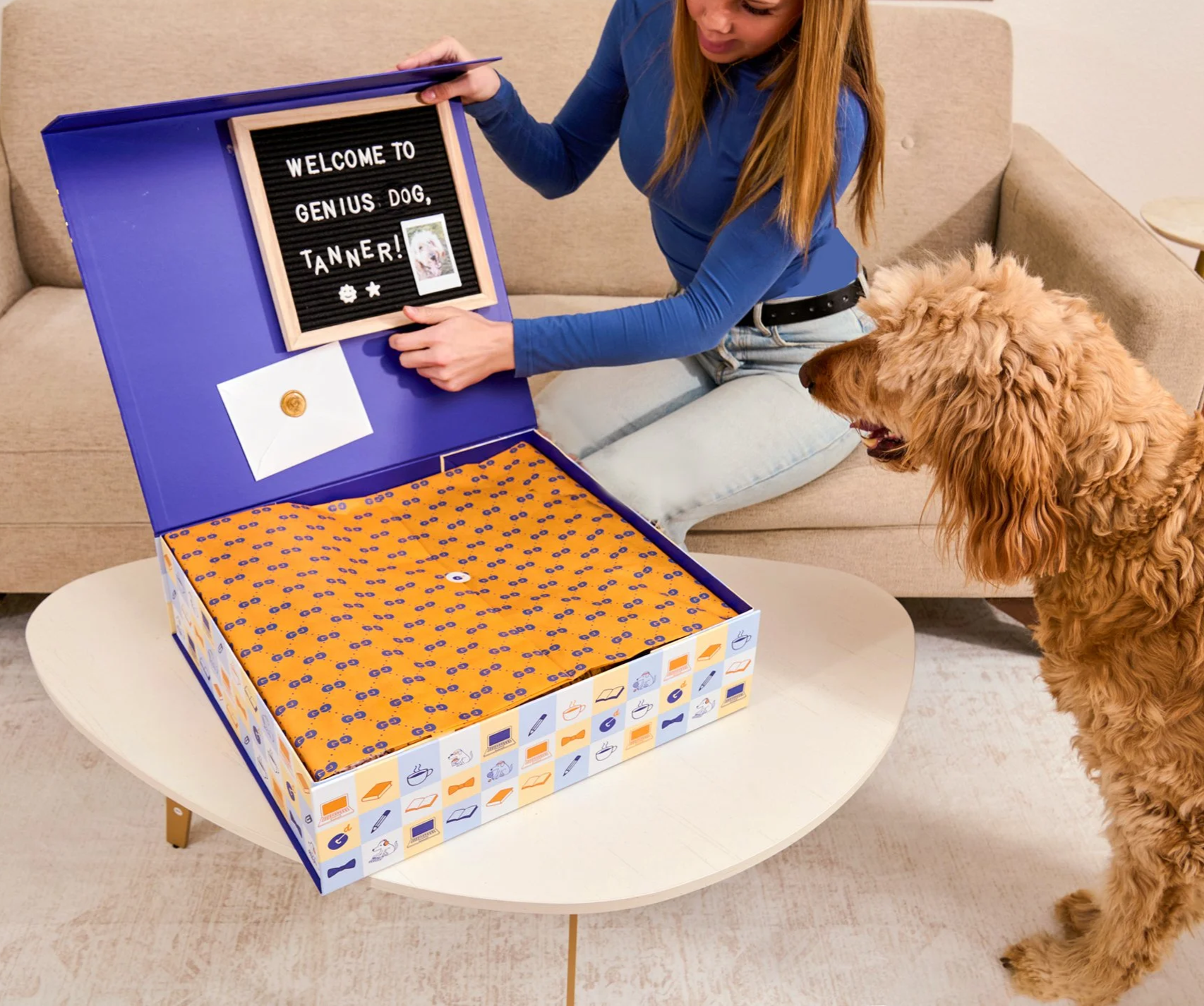 A woman opening a large box with a dog looking on. The box has a sign that says 'Welcome to Genius Dog TANNER!' and an envelope with a wax seal. The box is decorated with various icons related to dogs and toys.