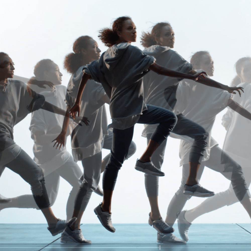 Multiple women in casual clothing performing synchronized dance or exercise moves on an outdoor stage with a sky background.
