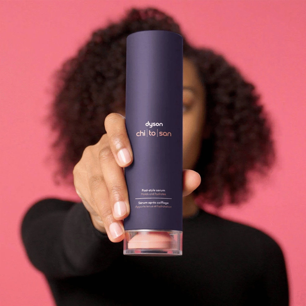 A person with curly hair holding a navy blue bottle of Dyson Chi-to-San post-style serum in front of a pink background.