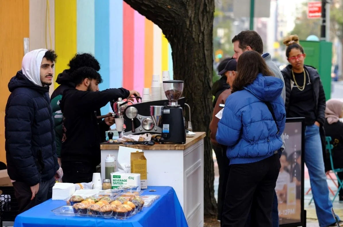 People ordering coffee from an outdoor stand with a colorful rainbow wall in the background.