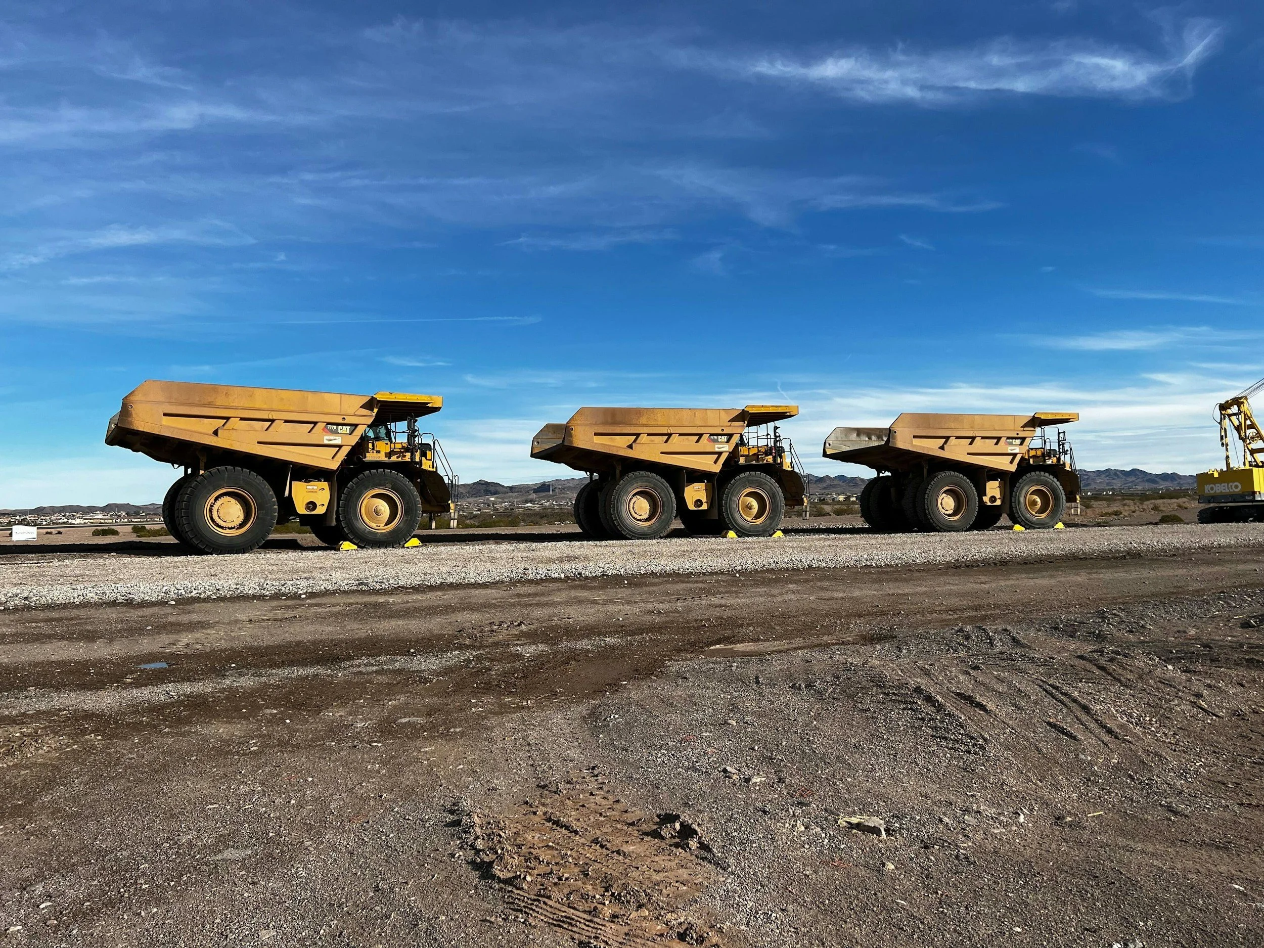 Dump trucks lined up on a desert construction site with mountains in the background under blue sky with wispy clouds.