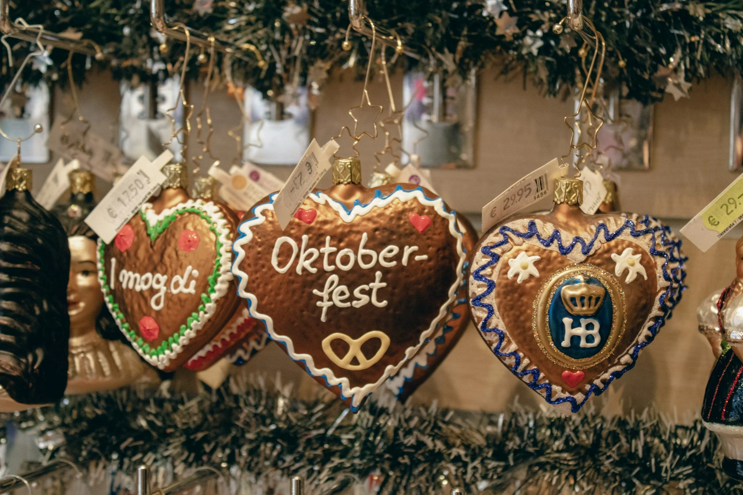Decorative heart-shaped cookies hanging from a Christmas tree branch, with inscriptions in German including 'Oktoberfest' and 'mögli', decorated with colorful icing and small edible embellishments, surrounded by tinsel.