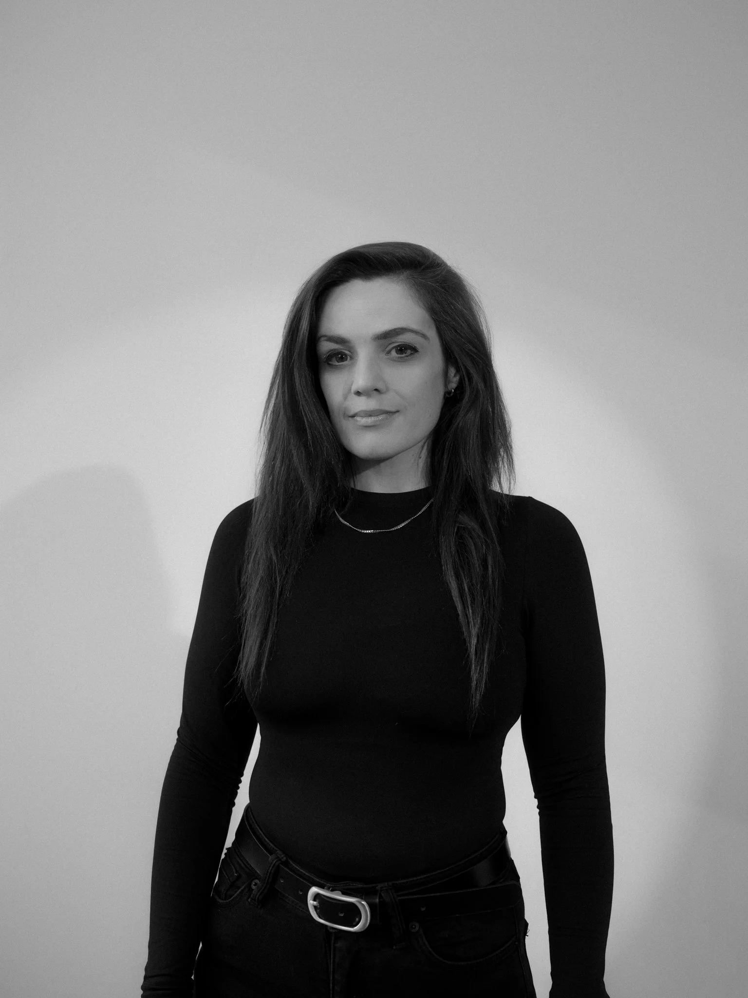 A woman with long dark hair, wearing a black long-sleeve top and jeans, standing against a plain light-colored background.