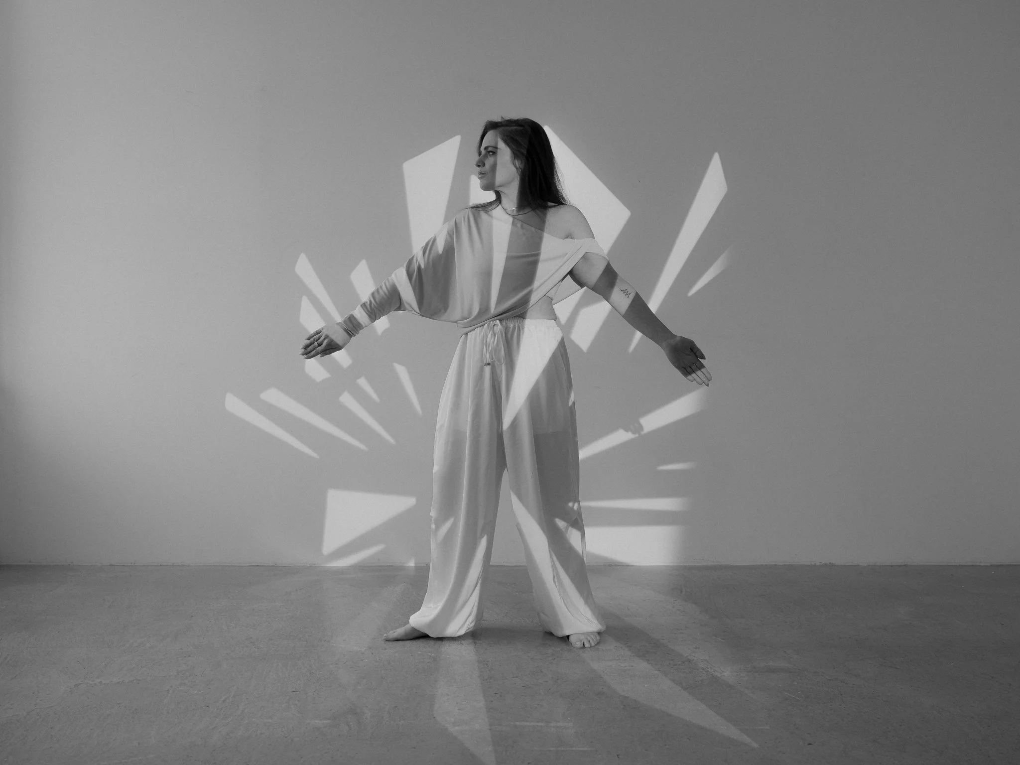 A woman dressed in a light-colored outfit with open shoulders stands in a gallery space with arms extended. Abstract geometric shapes are projected onto her and the plain wall behind her, creating a dynamic visual effect in black and white.