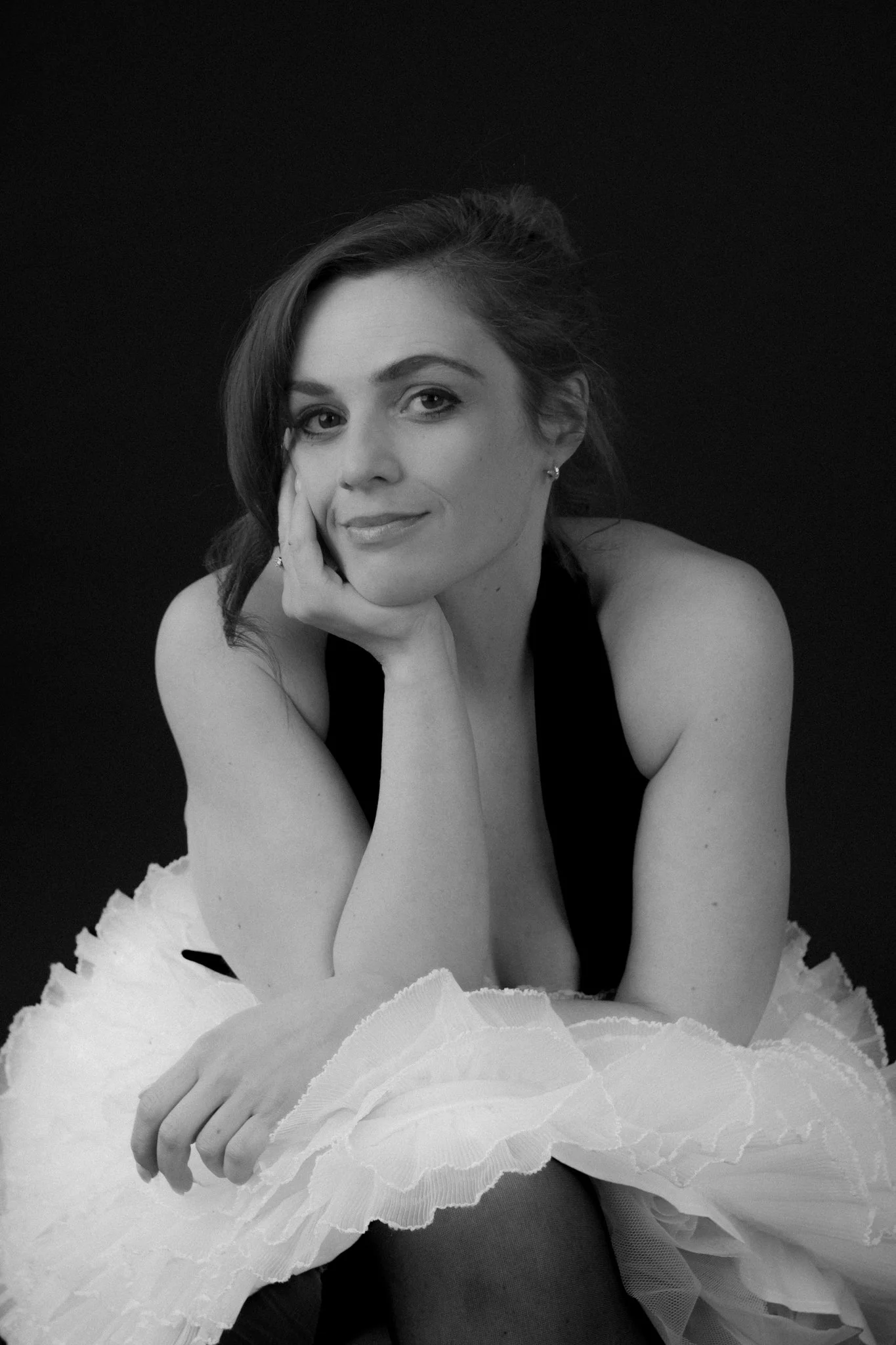 Black and white portrait of a woman with dark hair, resting her face on her hand, wearing a sleeveless top and a ruffled skirt, against a dark background.