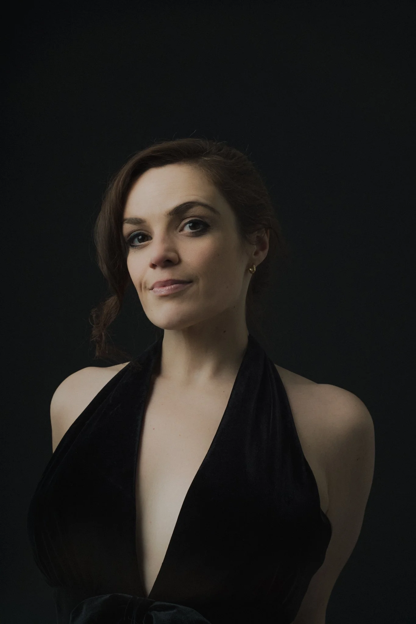 A woman with short brown hair, dressed in a black sleeveless top, standing against a black background.