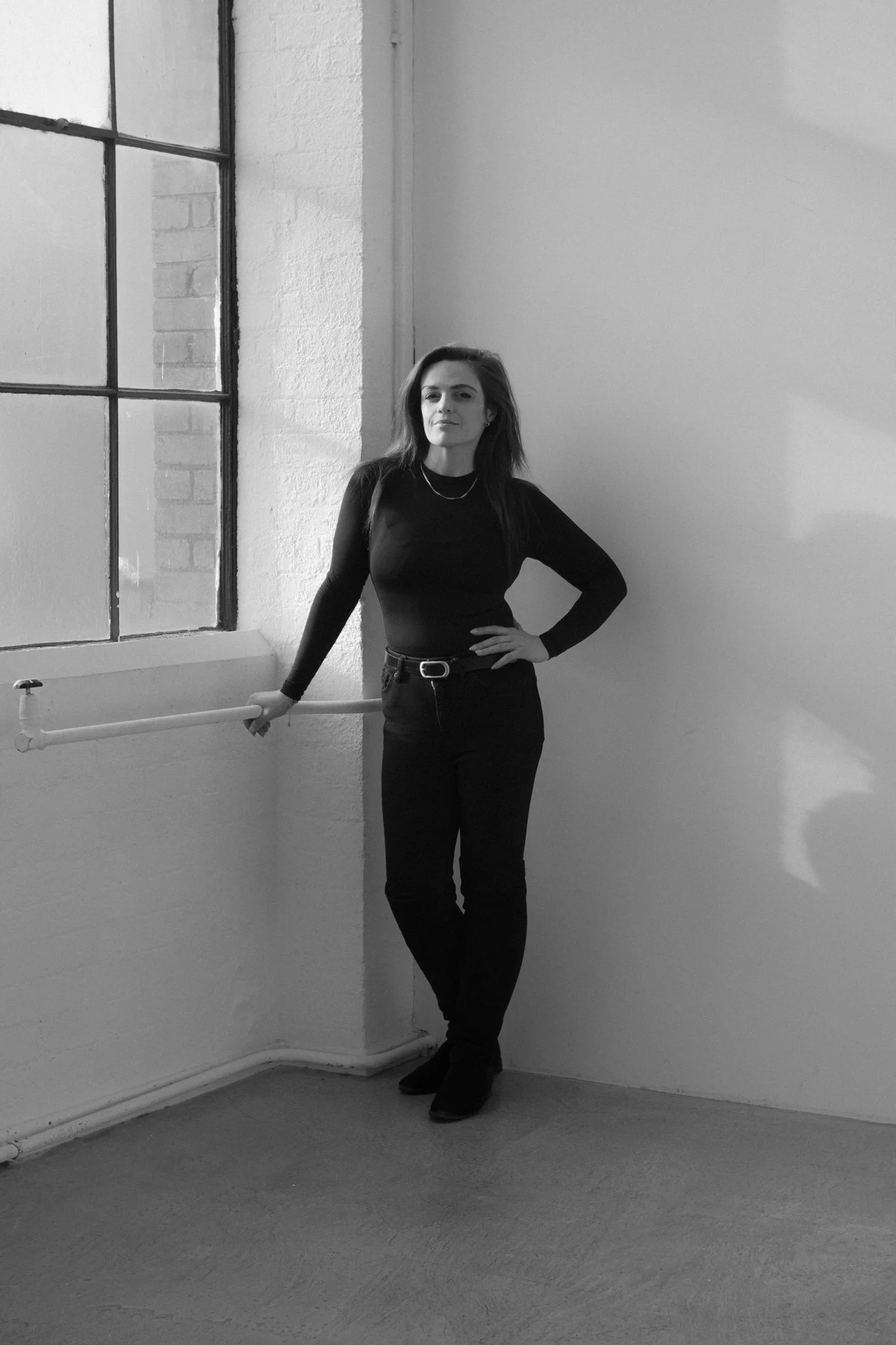 A woman standing in a room with a large window on the left, wearing a black long-sleeve shirt and black pants, posing with her right hand resting on a white horizontal bar attached to the wall.