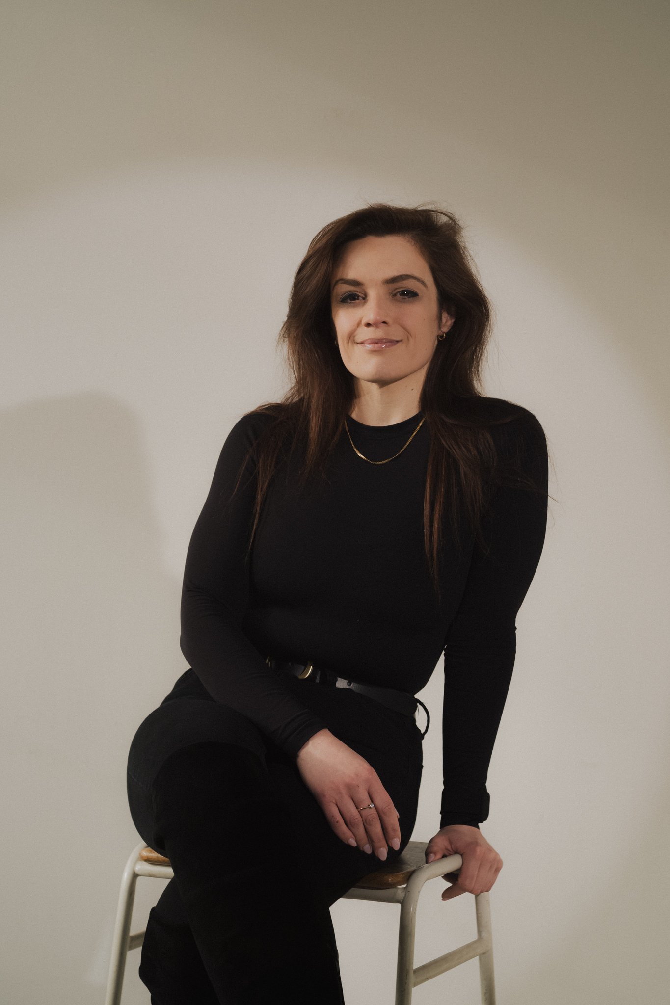 A woman with long brown hair, wearing a black long-sleeve top and black pants, sitting on a chair against a plain wall.