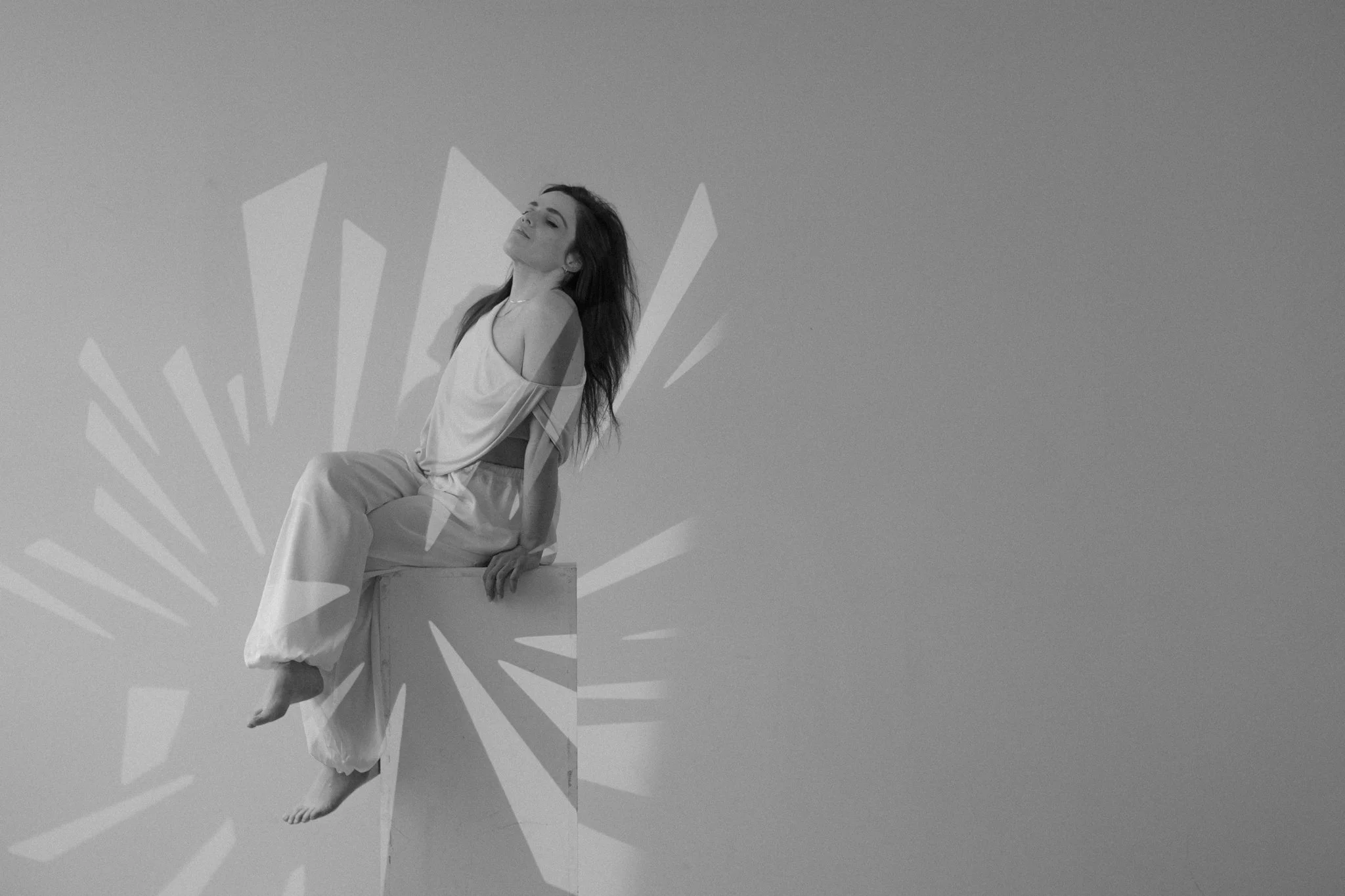 A woman sitting on a ledge with her eyes closed and head tilted back, posing against a plain background with graphic starburst lines.