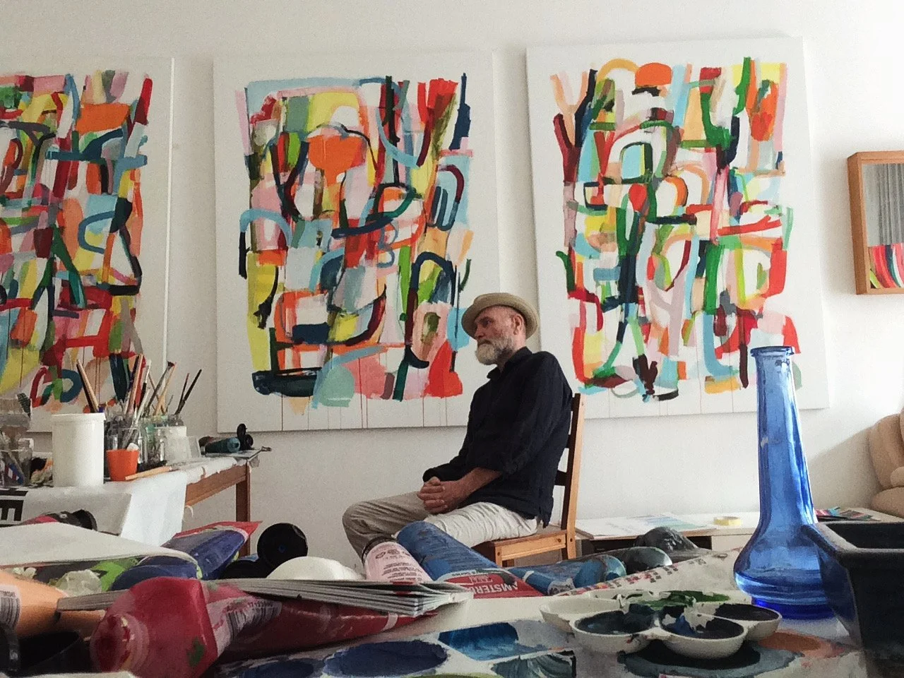 Artist sits in a wooden chair in front of his abstract, colorful art on the wall. Art supplies like paint and brushes fill the tables around him
