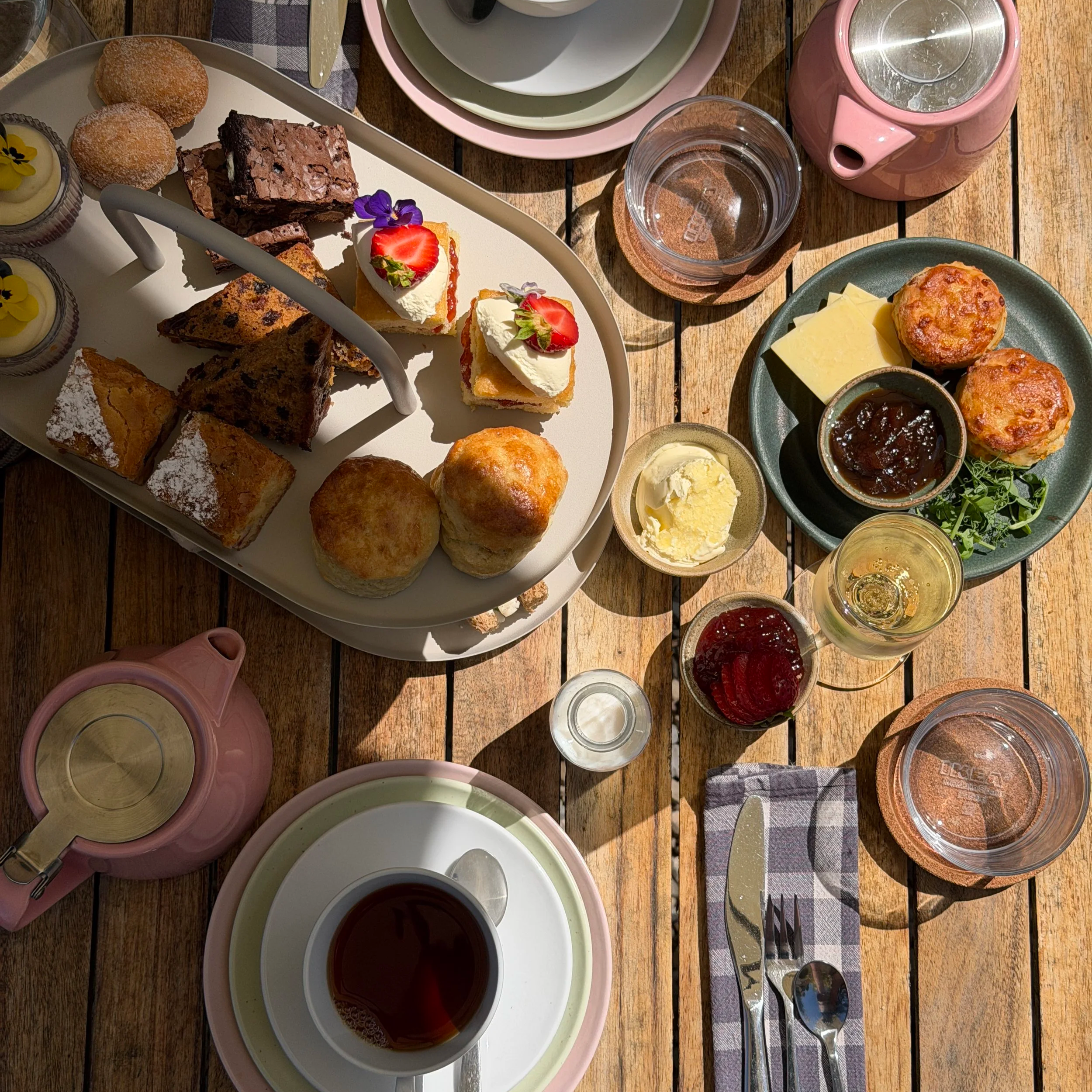 A white cup of tea on a saucer with a teaspoon, a pink teapot with a stainless steel insert on a wooden table, and scattered pink and yellow tulips.