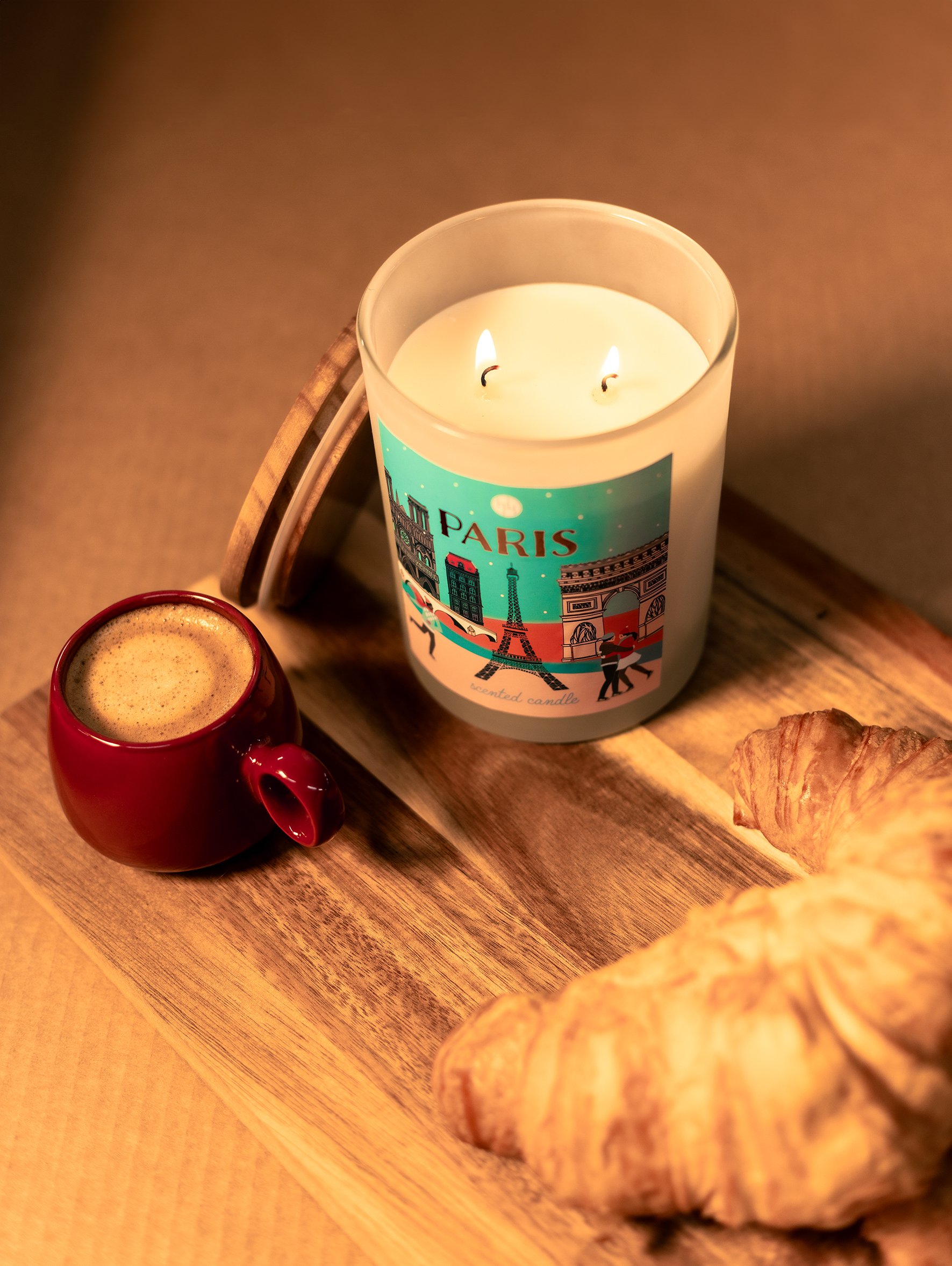 A wooden tray holding a red coffee cup with espresso, a lit candle in a glass jar labeled 'Paris,' and croissants on a beige surface.