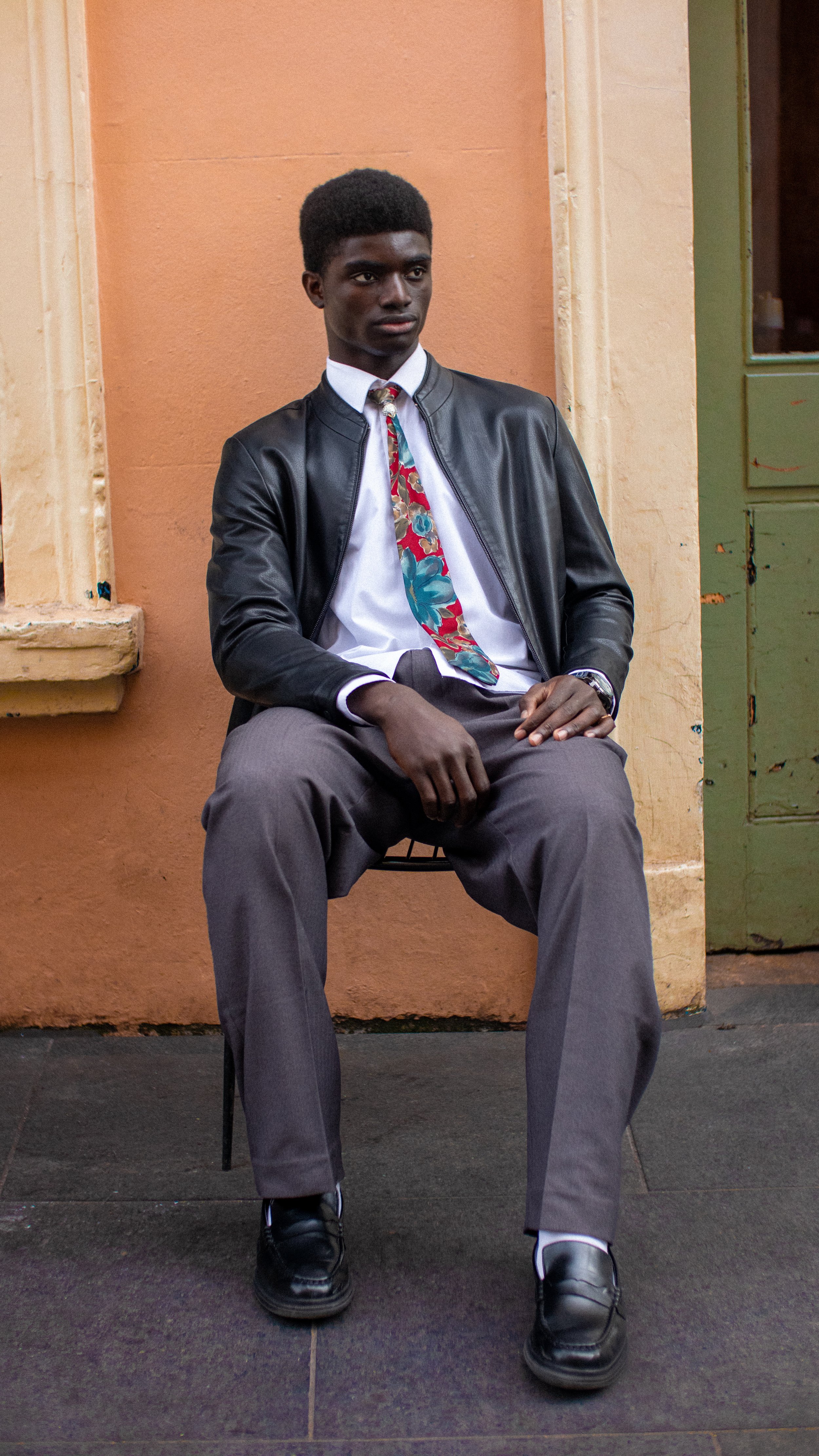 A person sitting against an orange wall, wearing a black leather jacket, white shirt, colorful tie, gray pants, and black shoes.