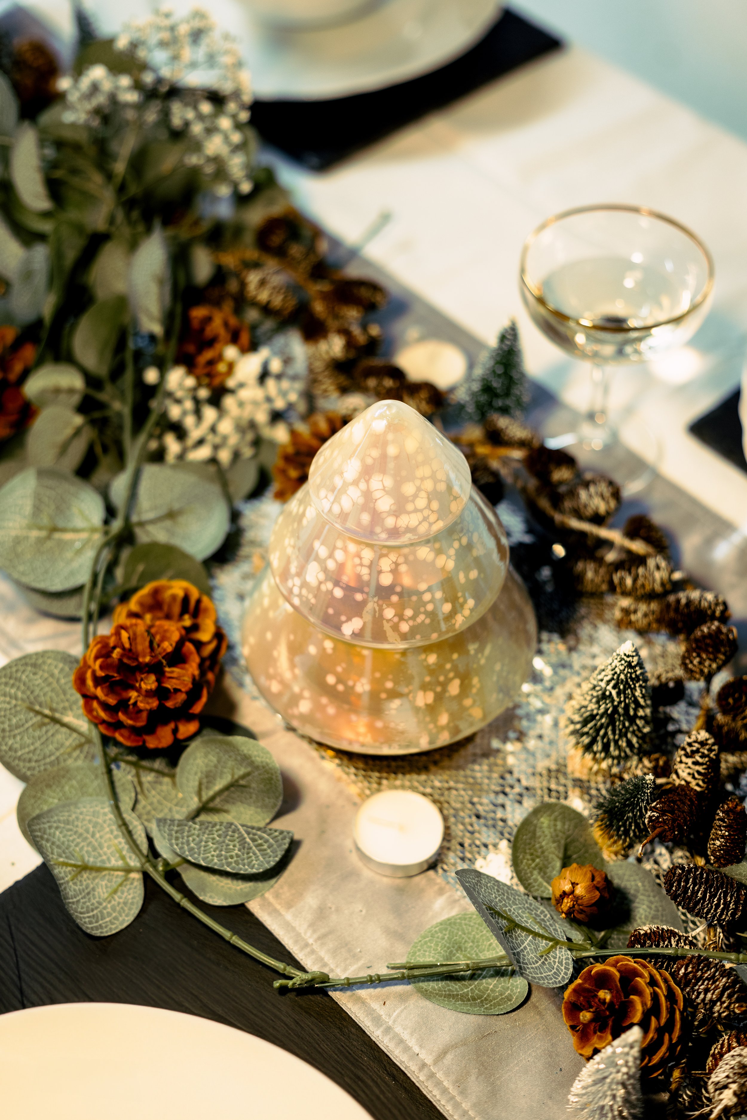 Festive table setting with decorative glass tree, pinecones, greenery, small candles, and a glass of white wine.